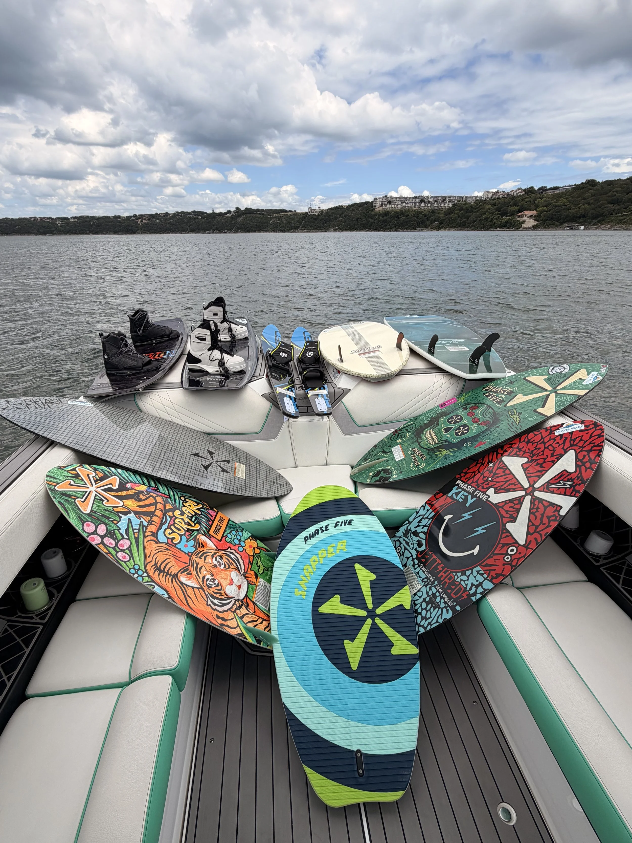 Boarding area of a boat with multiple colorful paddleboards and accessories on a padded seating area, water and distant hillside in the background.