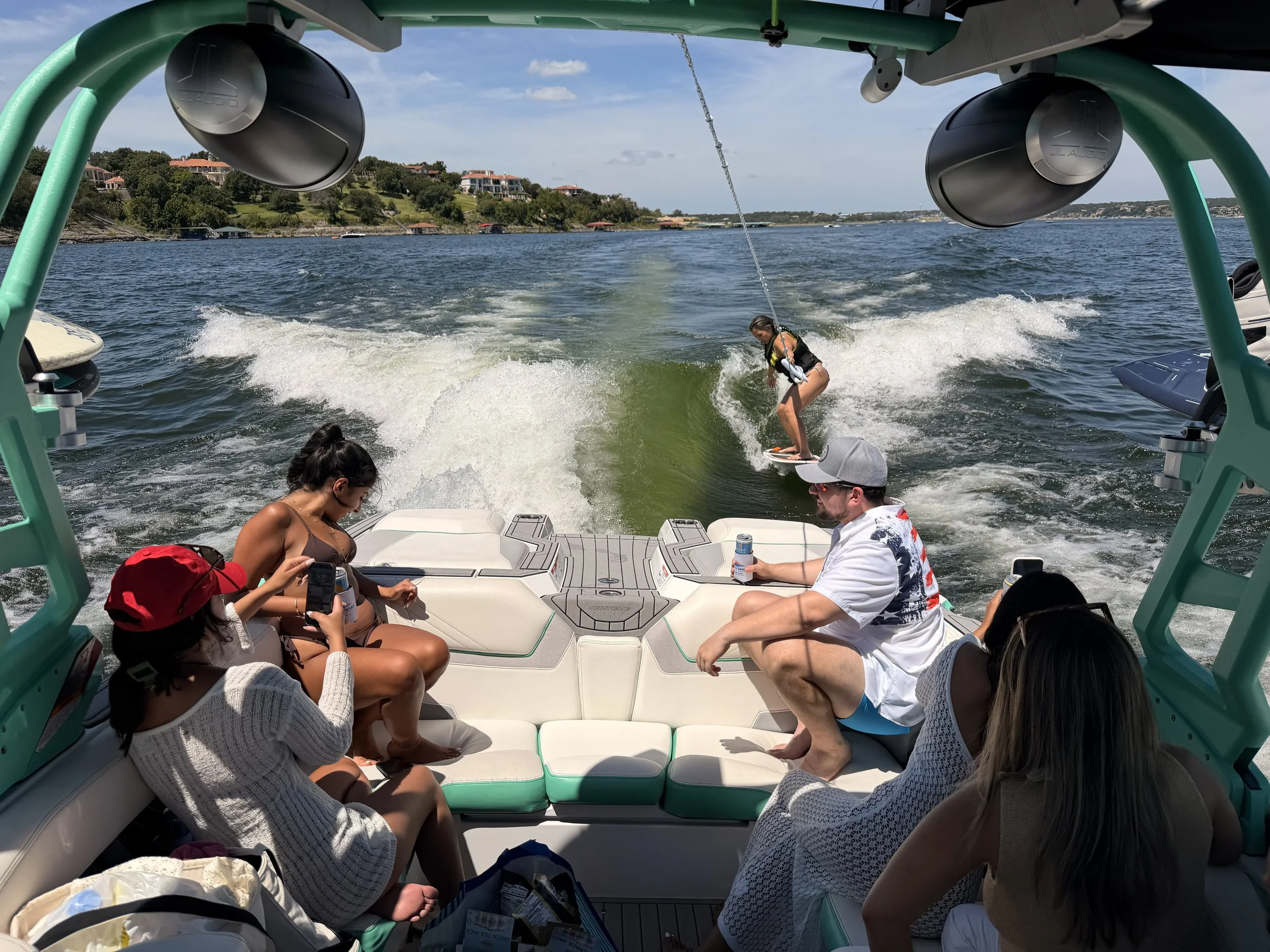Group of people enjoying a boat ride on a lake with a hillside residential area in the background, some are sitting while one person attempts a wakeboard stunt at the back of the boat.