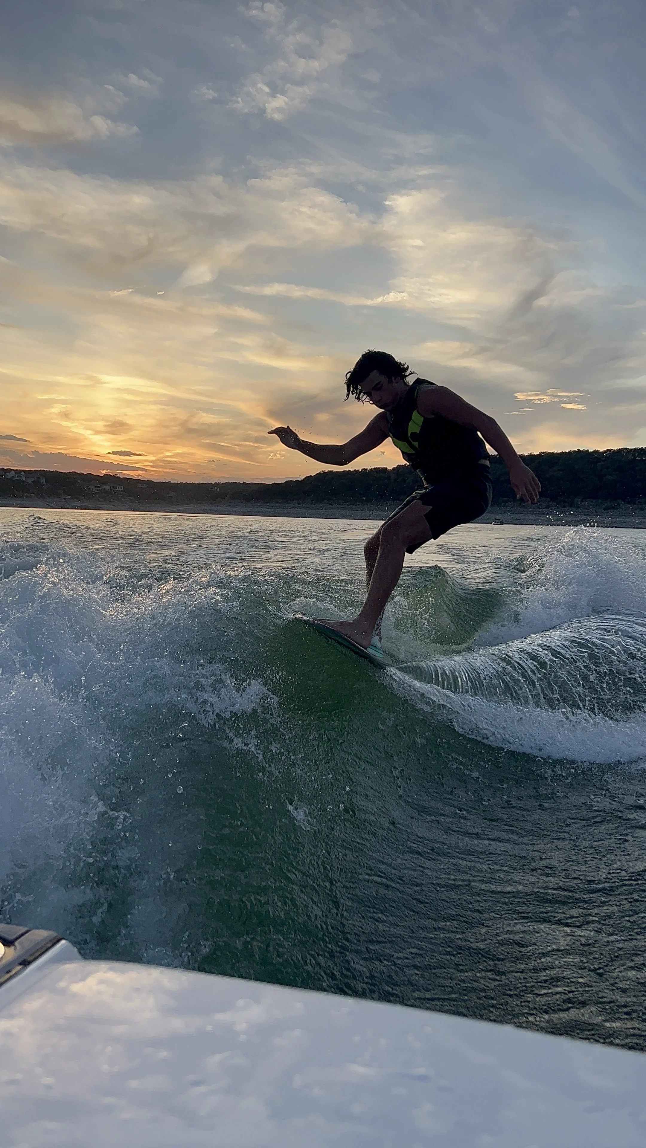A person surfing on a wave during sunset with a partly cloudy sky and a distant shoreline.