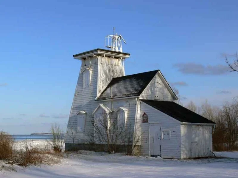 An old white lighthouse with a small attached building, set in a snowy landscape with a clear blue sky.