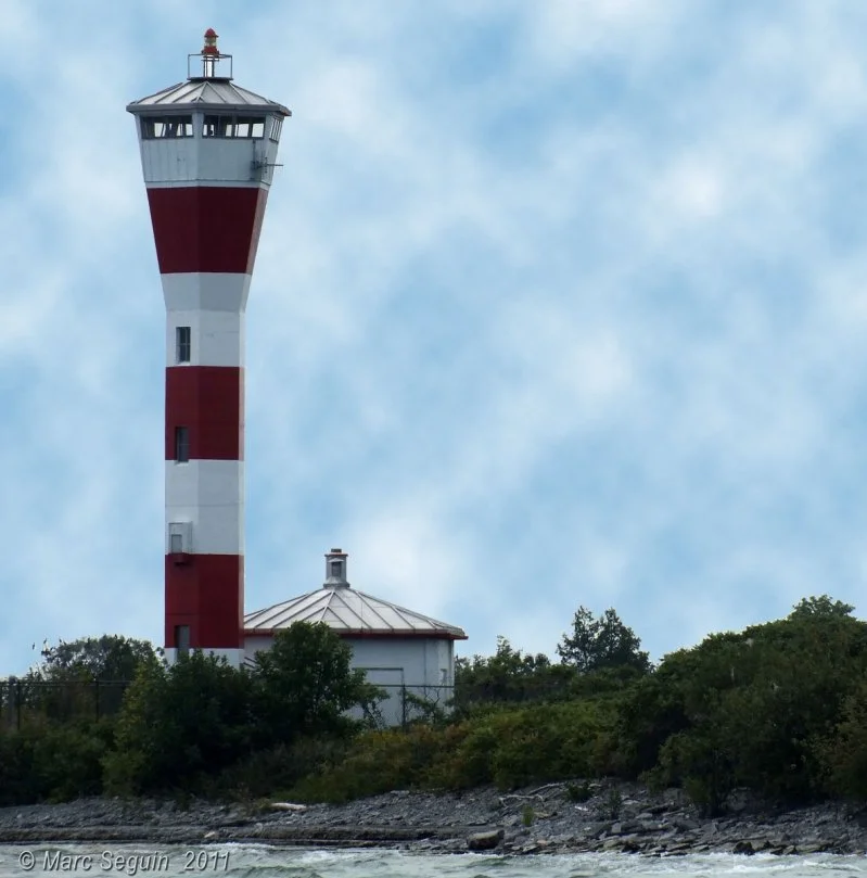 A red and white striped lighthouse on a grassy hill near water with a partly cloudy sky in the background.