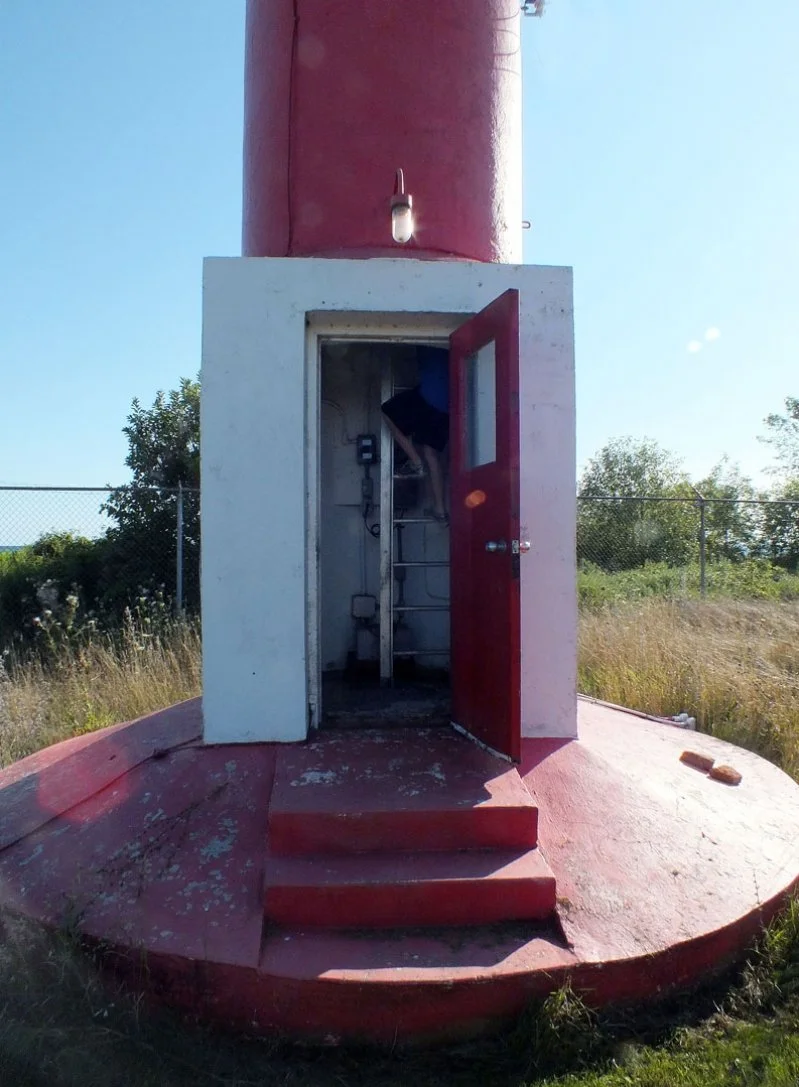 View of a small lighthouse with a red and white exterior, an open door revealing a ladder inside, and surrounded by grass and trees under a clear blue sky.
