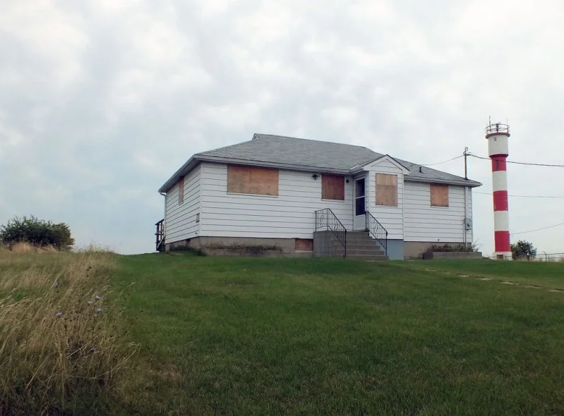 An abandoned white house with boarded-up windows on a grassy hill, with a red and white striped lighthouse in the background.
