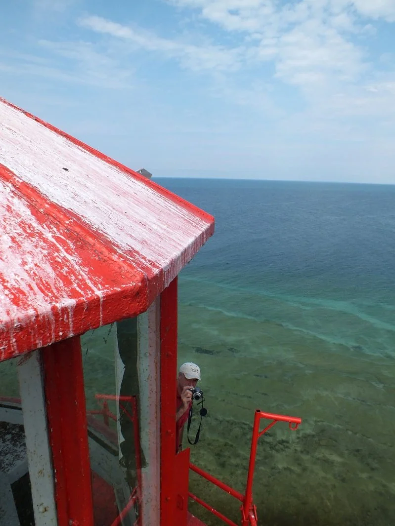 A person taking a photo from a red and white lighthouse or observation tower overlooking the ocean. The sky is partly cloudy and the water is clear with visible underwater features.