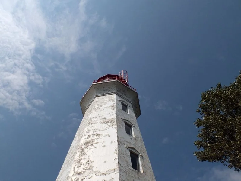 A white lighthouse with a red top against a partly cloudy blue sky, with a tree on the right side.