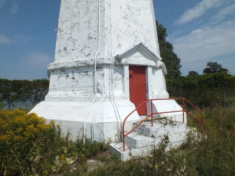 A white lighthouse with a red door and steps, surrounded by overgrown bushes under a partly cloudy sky.