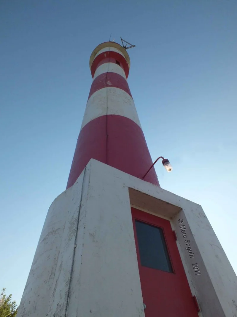 Low-angle view of a red and white striped lighthouse with a small window and a light fixture, set against a clear blue sky.
