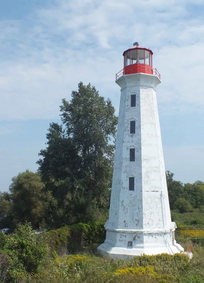 A tall, white lighthouse with a red top situated among greenery and trees, under a partly cloudy sky.