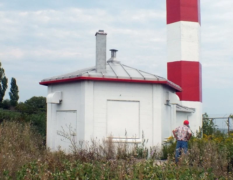 A white utility building with a metal roof and a chimney, beside a tall red-and-white striped smokestack, and a man wearing a red cap and plaid shirt standing nearby in overgrown grass.