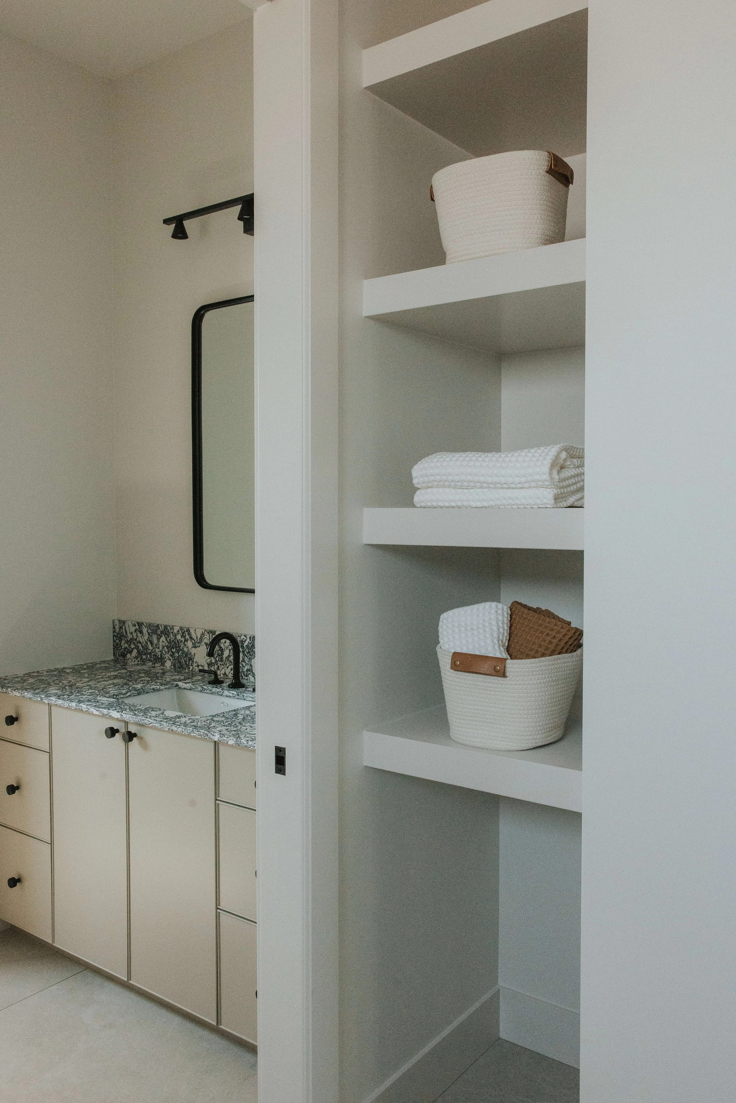 A modern kitchen with beige cabinets, a marble countertop, a black faucet, and a small mirror on the wall. Next to the kitchen area is a built-in white shelving unit with three shelves, holding a woven basket and folded textiles.