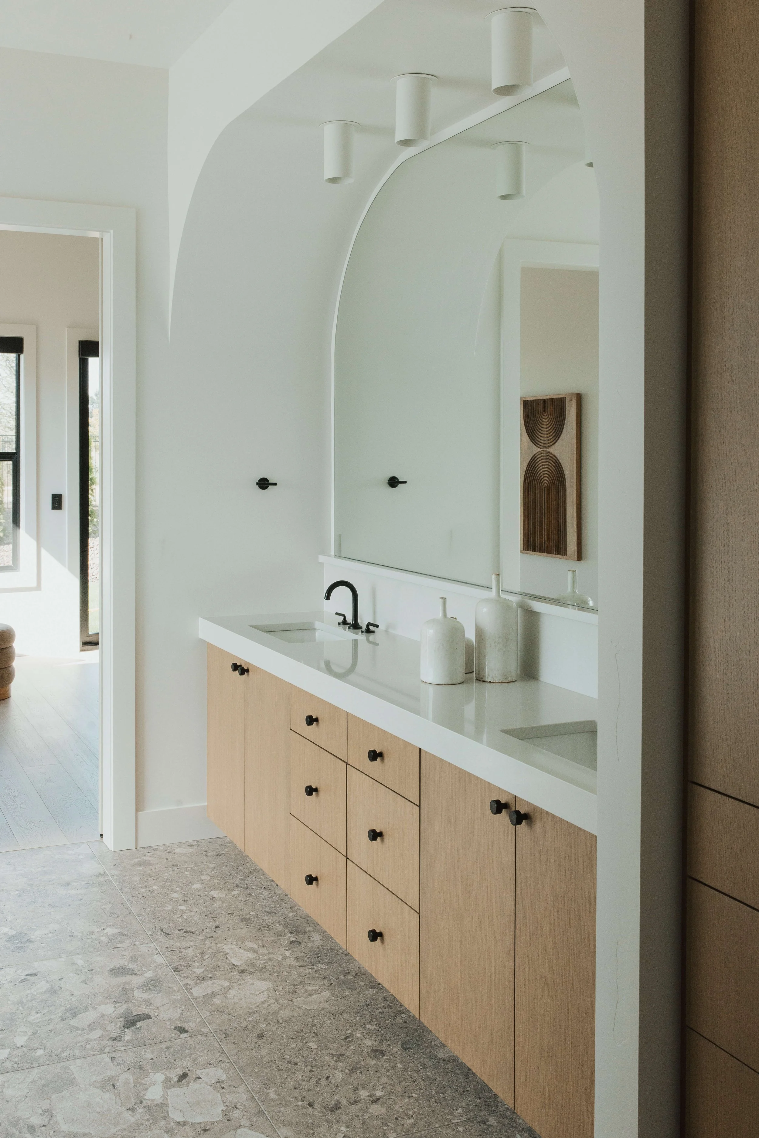 Modern kitchen with light wood cabinets, white countertop, black handles, and a large mirror. Decor includes white vases. The floor is made of large gray tiles. There is a window with black trim in the background.