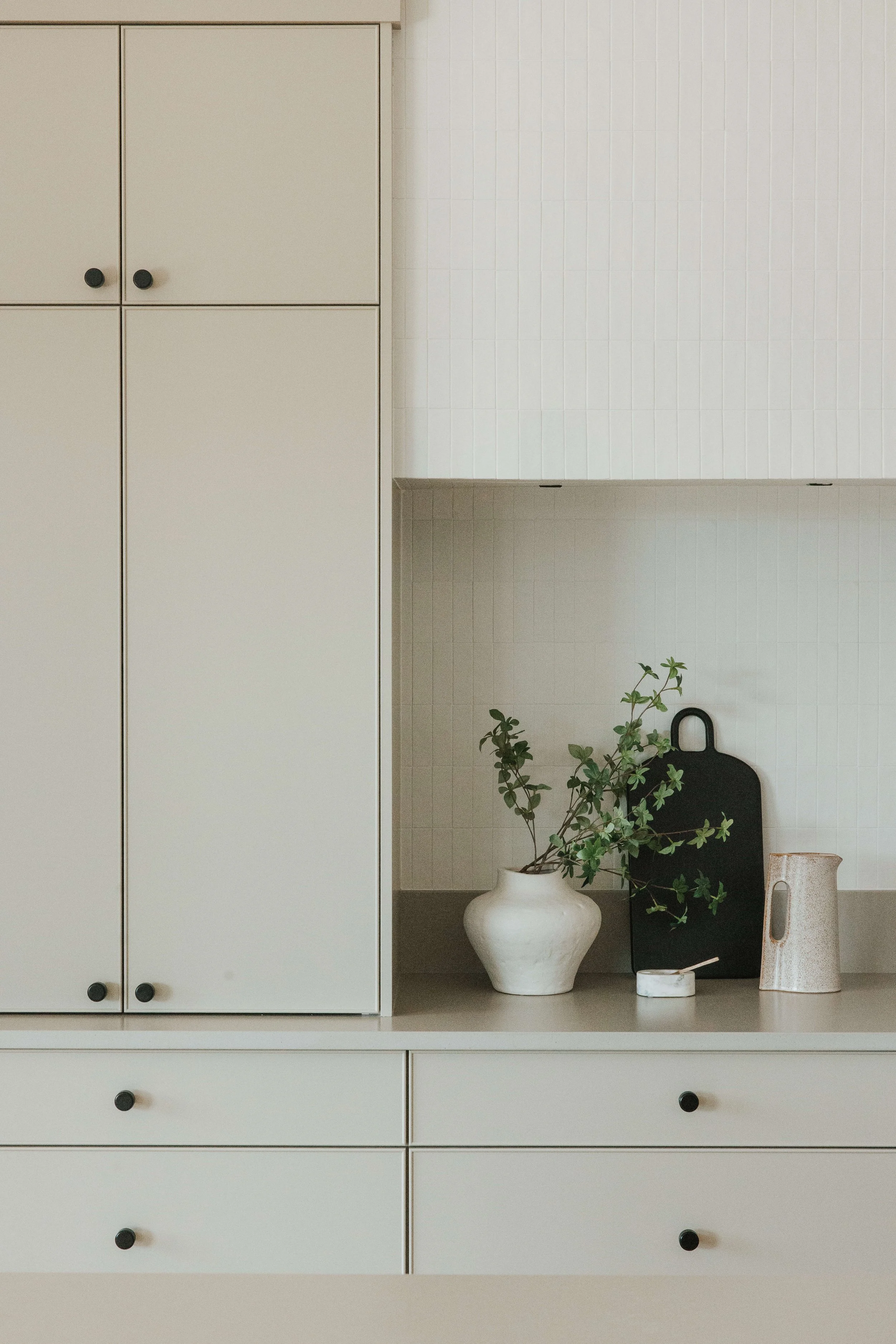 A white cabinet with black knobs and a set of drawers, beside a shelf with a potted green plant in a white vase, a small black object, and decorative vases on a light-colored countertop, against a white tiled wall.