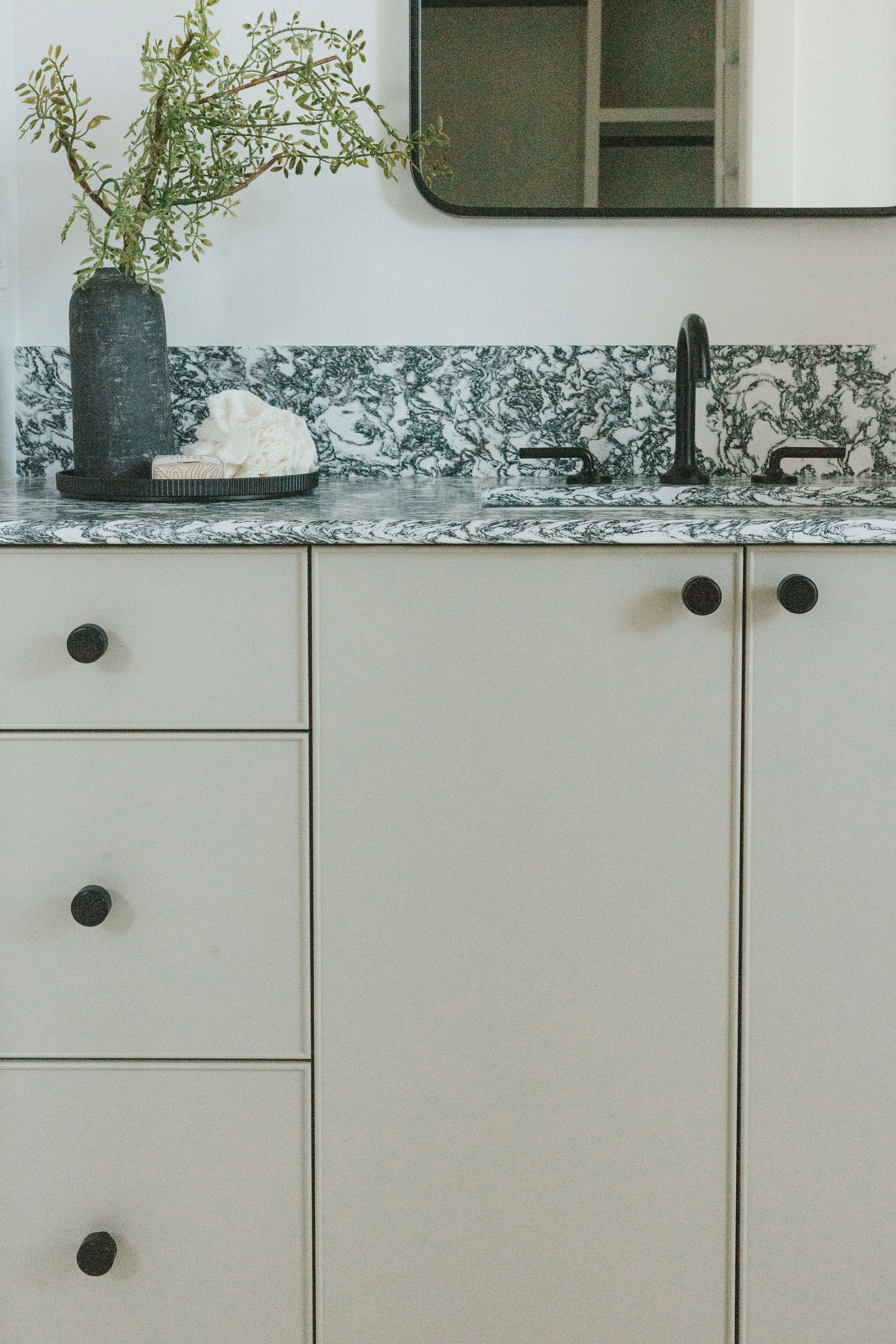 A kitchen countertop with a black vase holding a green leafy plant, a soap dish with a bar of soap, and a soap saver. There is a black sink with a black faucet and a large mirror above the countertop.