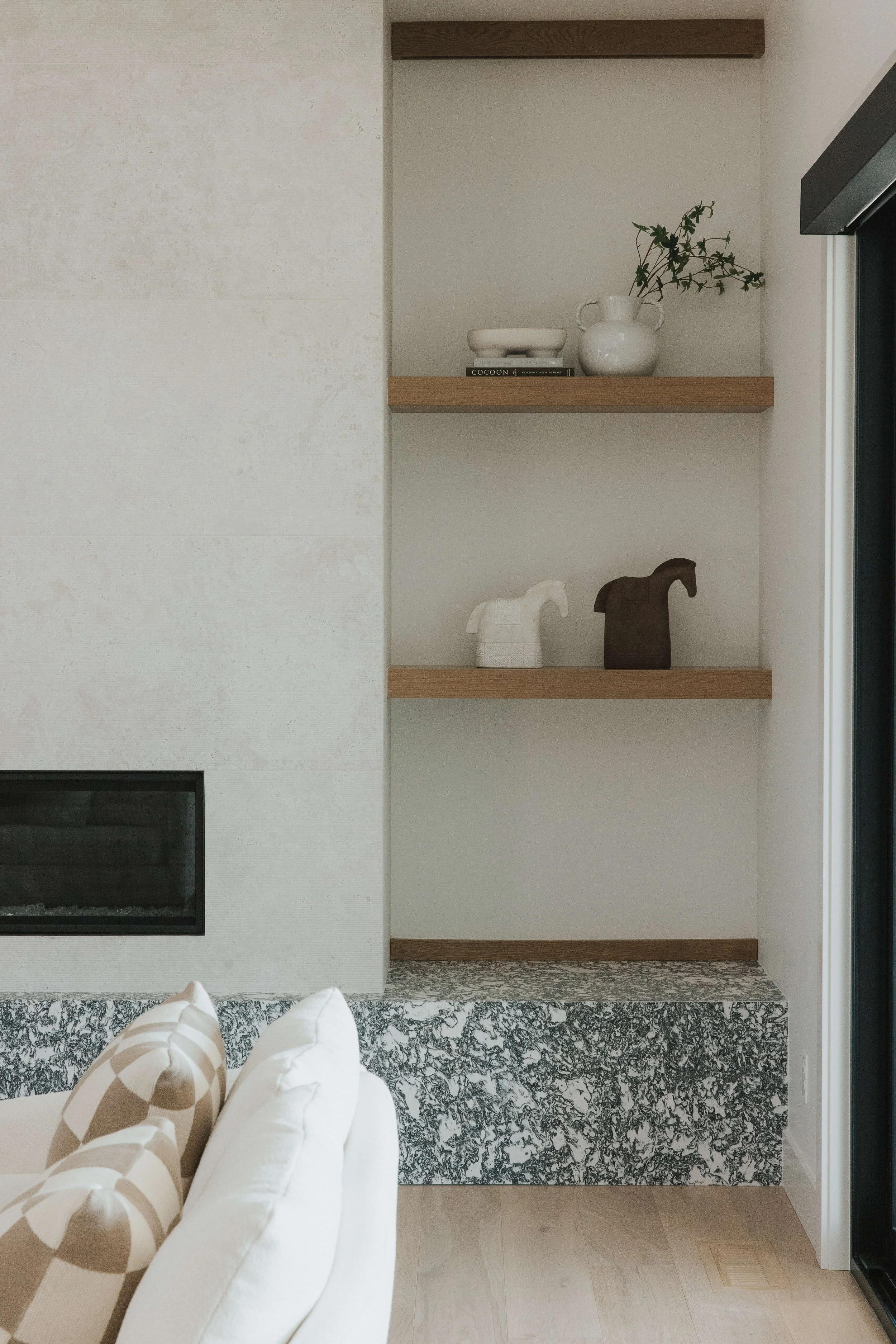 Living room with white sofa, beige and white patterned pillows, a fireplace, and built-in wooden shelves with decorative items including a white vase with greenery, a shallow bowl, and small horse sculptures, with a patterned rug on the floor.