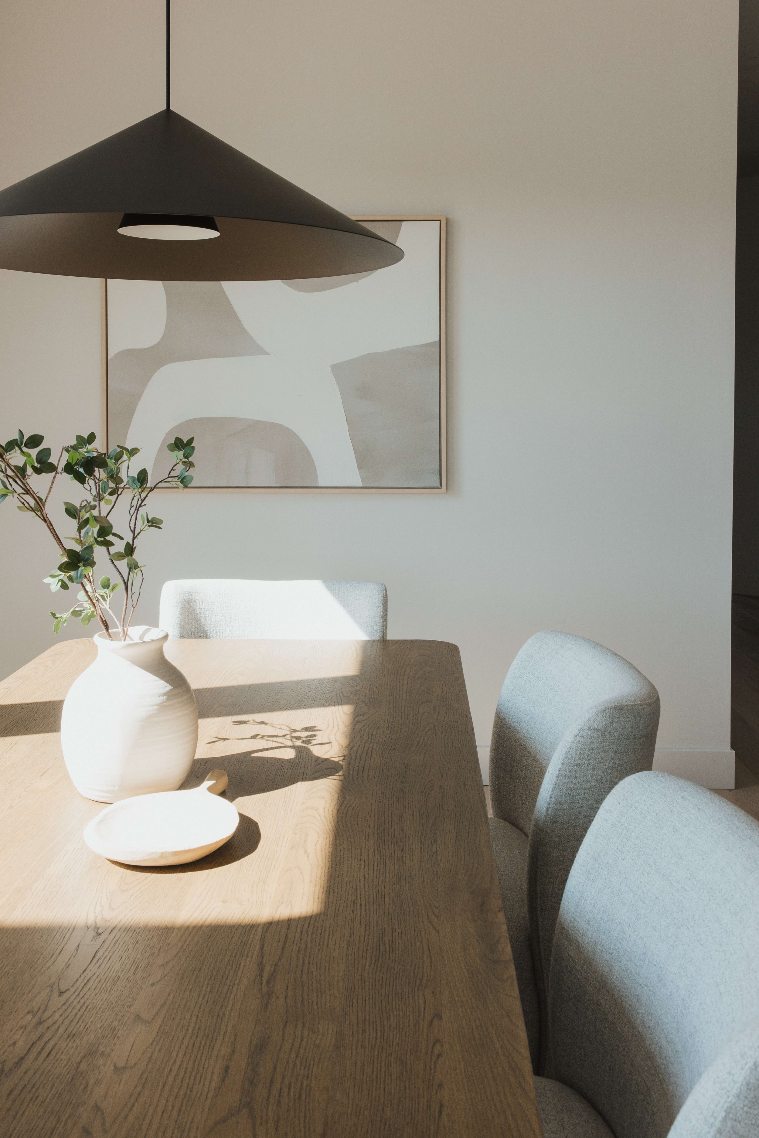 A dining room with a wooden table, gray upholstered chairs, a white ceramic vase with green foliage, and abstract artwork on a off-white wall. A black pendant light hangs above the table.