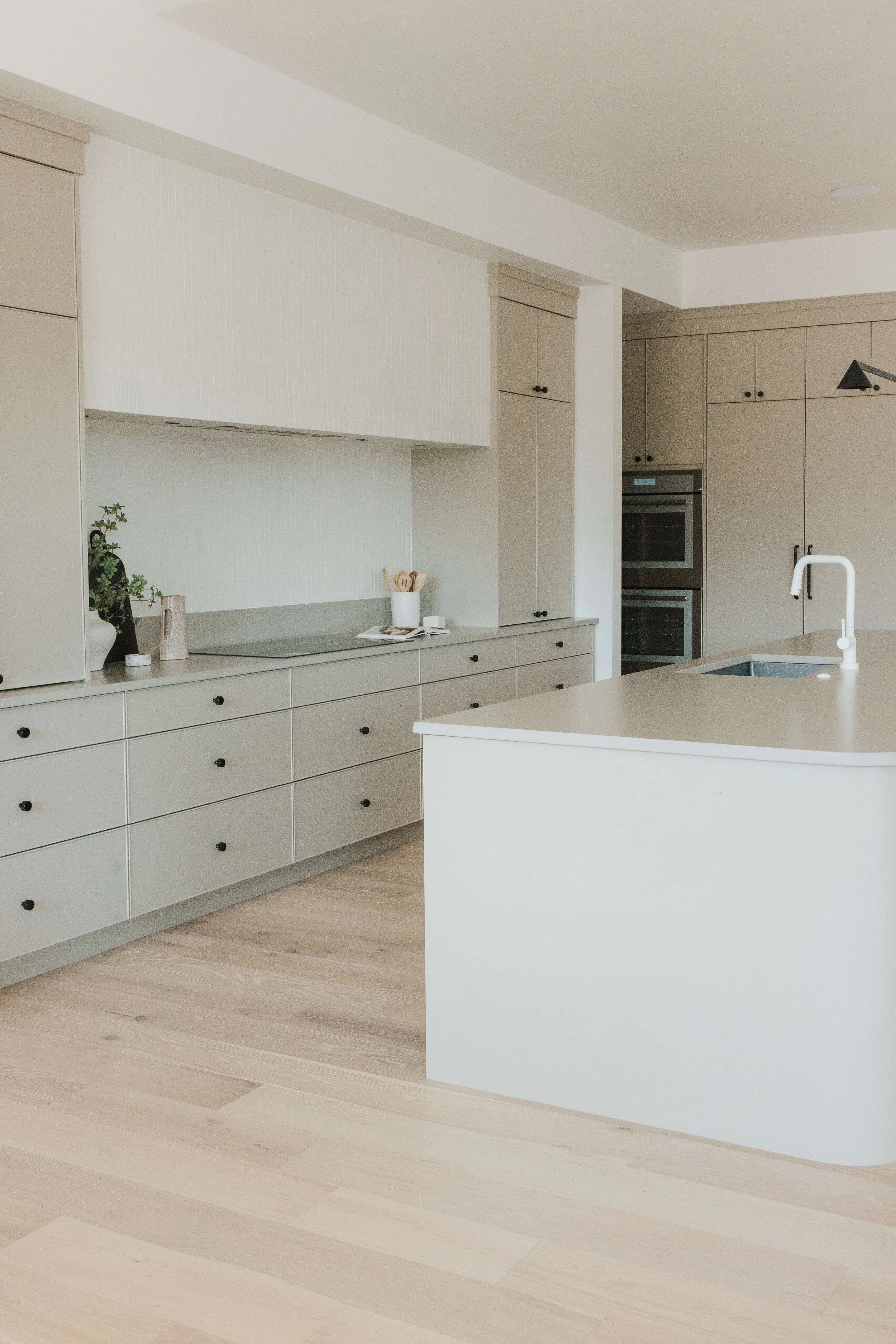 Modern kitchen with beige cabinetry, white walls, light wood flooring, a kitchen island with a white sink and faucet, and decorative items on the counter.