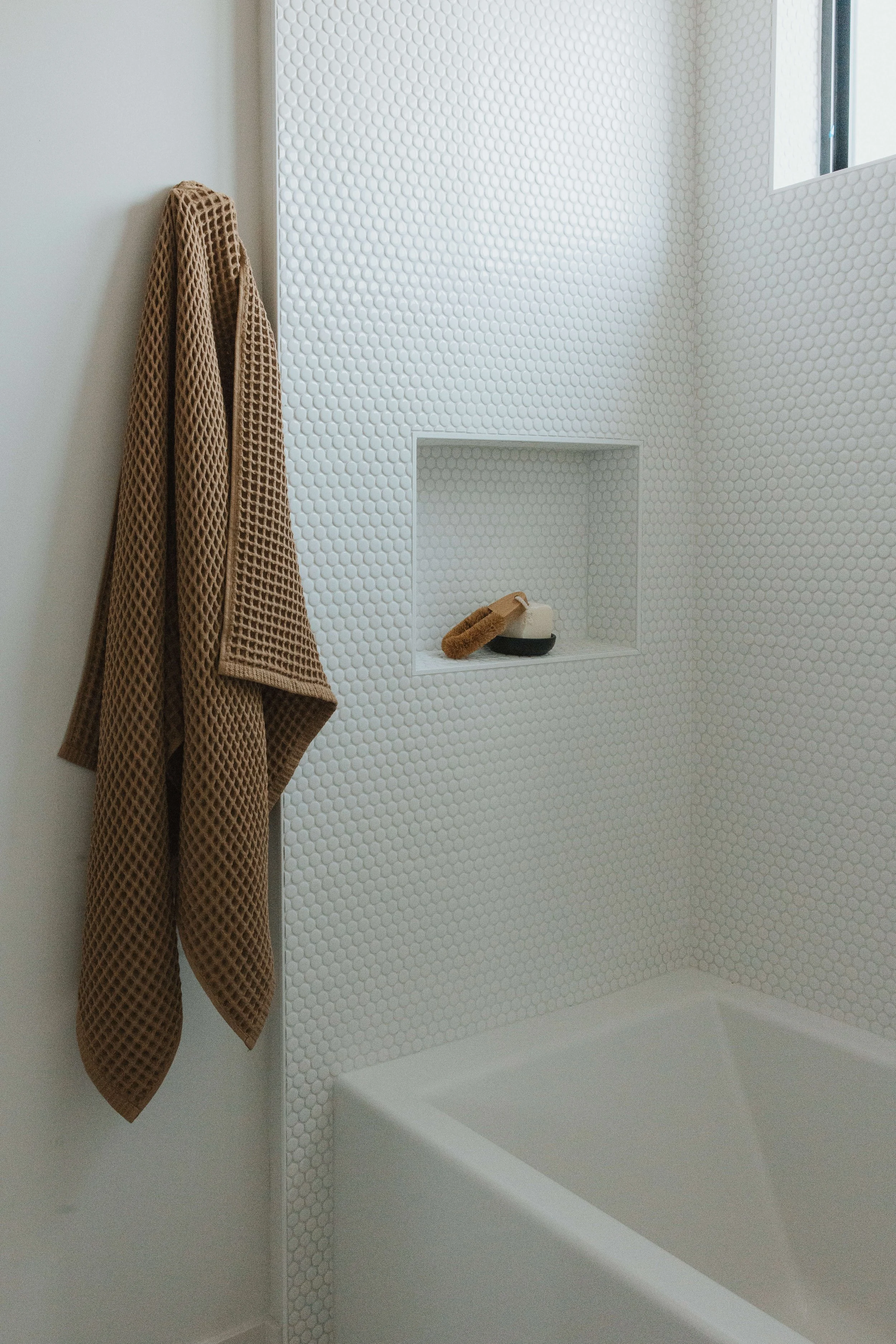 A shower with white textured wall tiles, a small built-in shelf holding a natural brush and clay bowl, and a brown washcloth hanging on a hook.