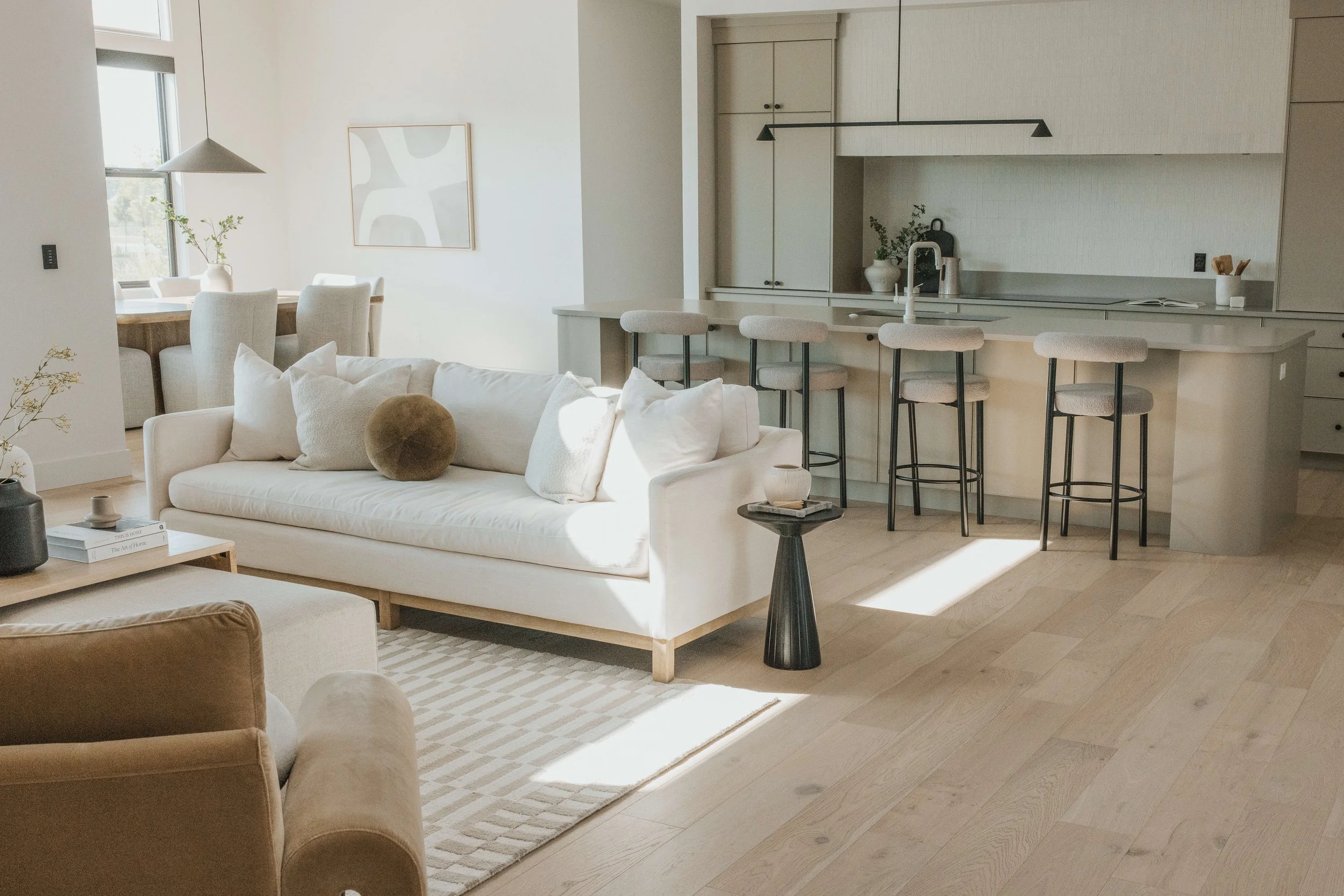 A bright, modern living space with a white sofa, beige cushions, and a black side table. A dining area with white upholstered chairs around a wooden table is visible in the background near a window with natural light. The open kitchen features white 