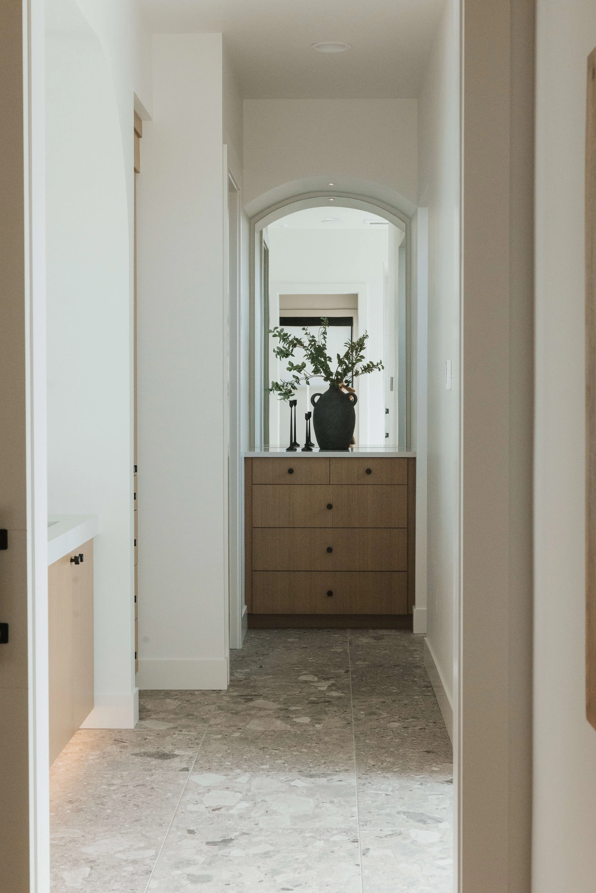 A narrow hallway with light-colored walls, a tiled floor, and a wooden dresser at the end. On top of the dresser are a large black vase with green branches and three black candlesticks. An arched doorway leads to another room with a fireplace and win