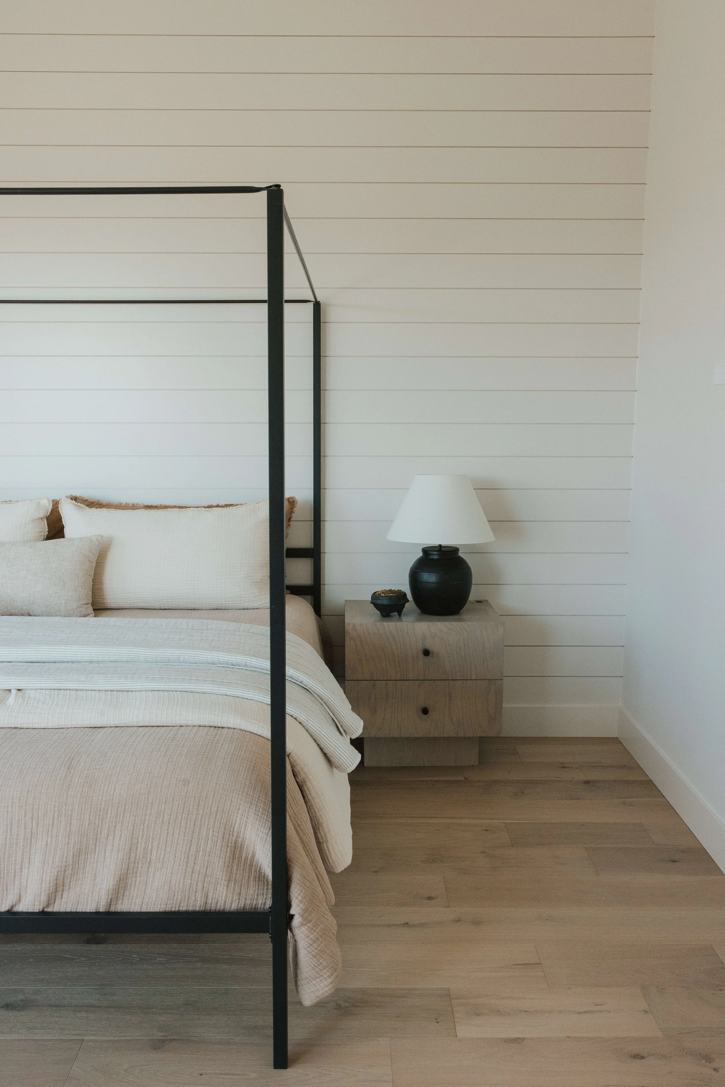 A bedroom with a minimalist design, featuring a bed with beige bedding and pillows, a wooden nightstand with a black and white lamp, and a white wall with subtle horizontal lines.