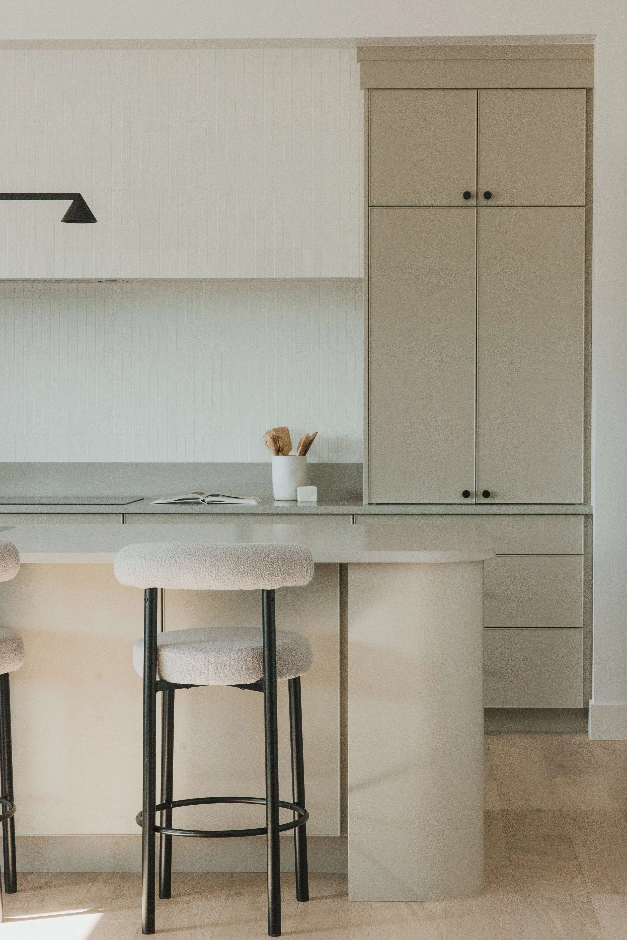 Modern kitchen with beige cabinetry, a small countertop with an open book and a white mug containing wooden utensils, a white stool, and a black metal barstool, with light-colored wooden flooring.