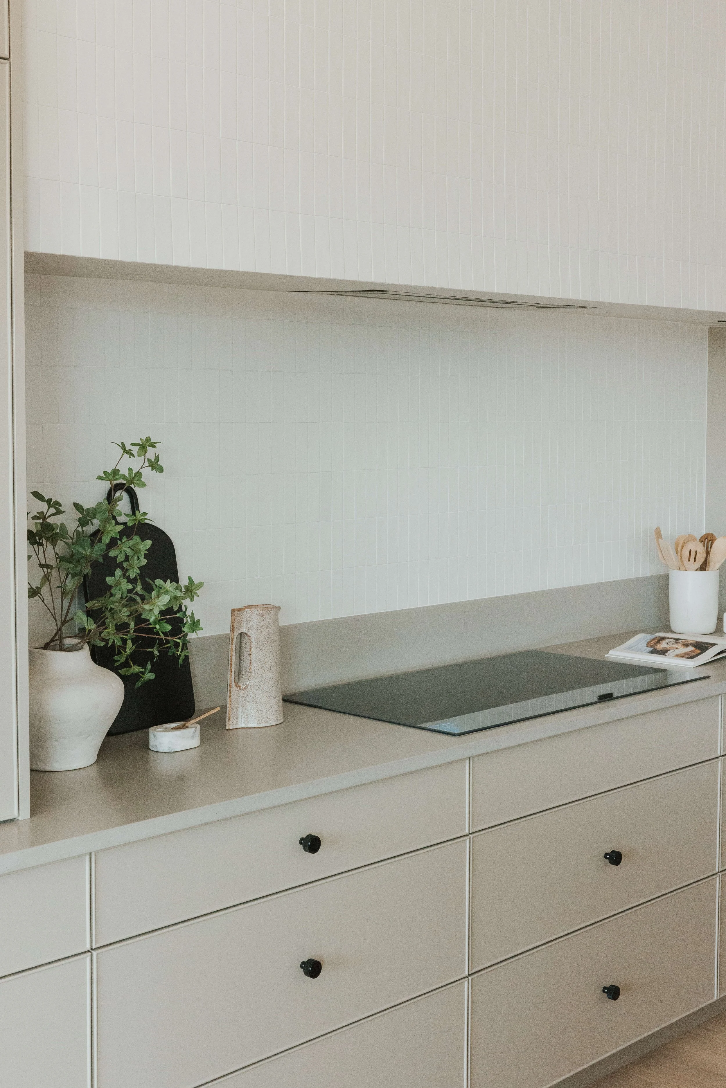 A kitchen countertop with a potted plant, a white ceramic mug, a candle, a small dish, and a recipe book. Behind is a white tiled backsplash and beige cabinets with black knobs.