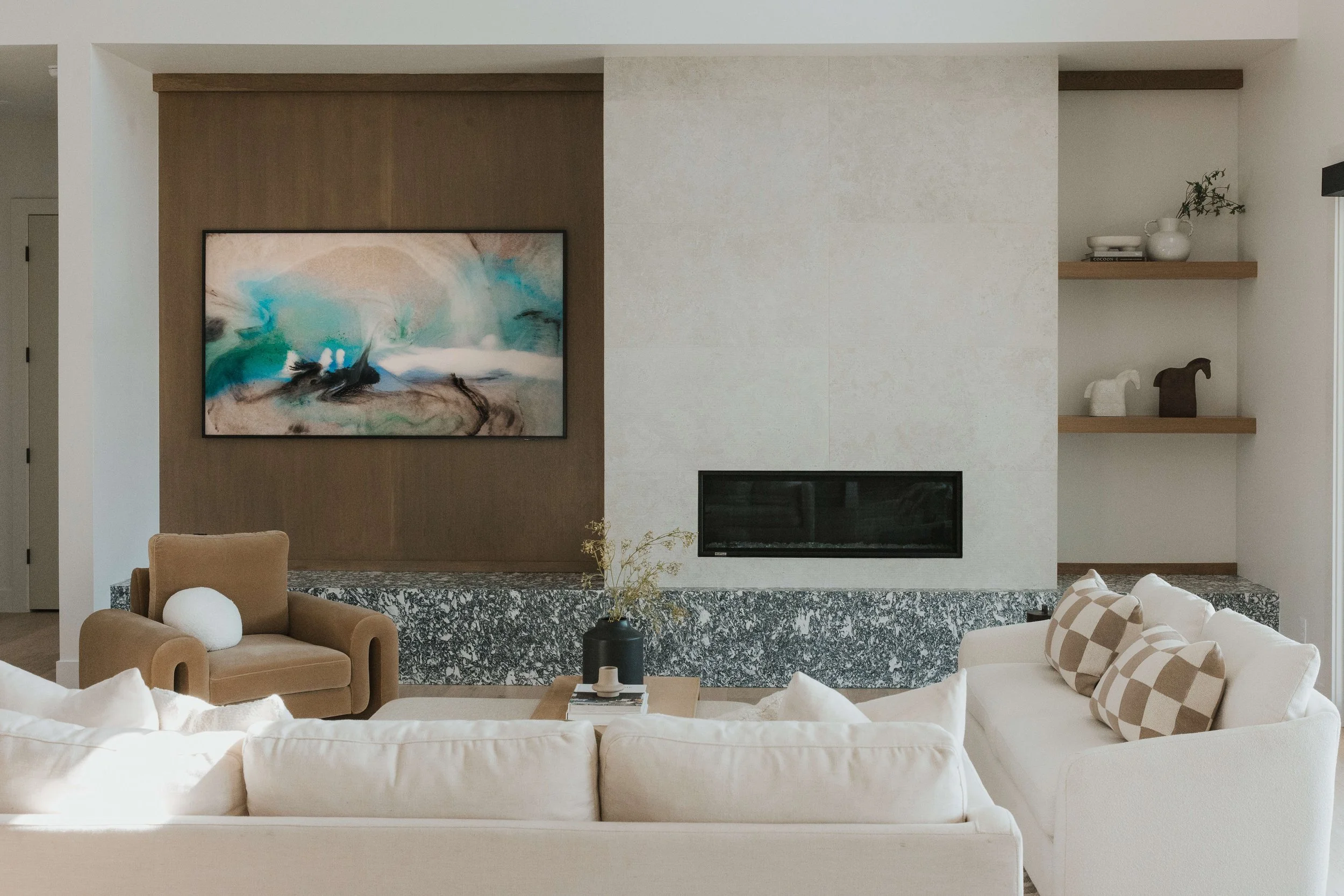 Modern living room with white sofas, beige armchair, wall-mounted abstract art, fireplace, and built-in shelves with decorative items.