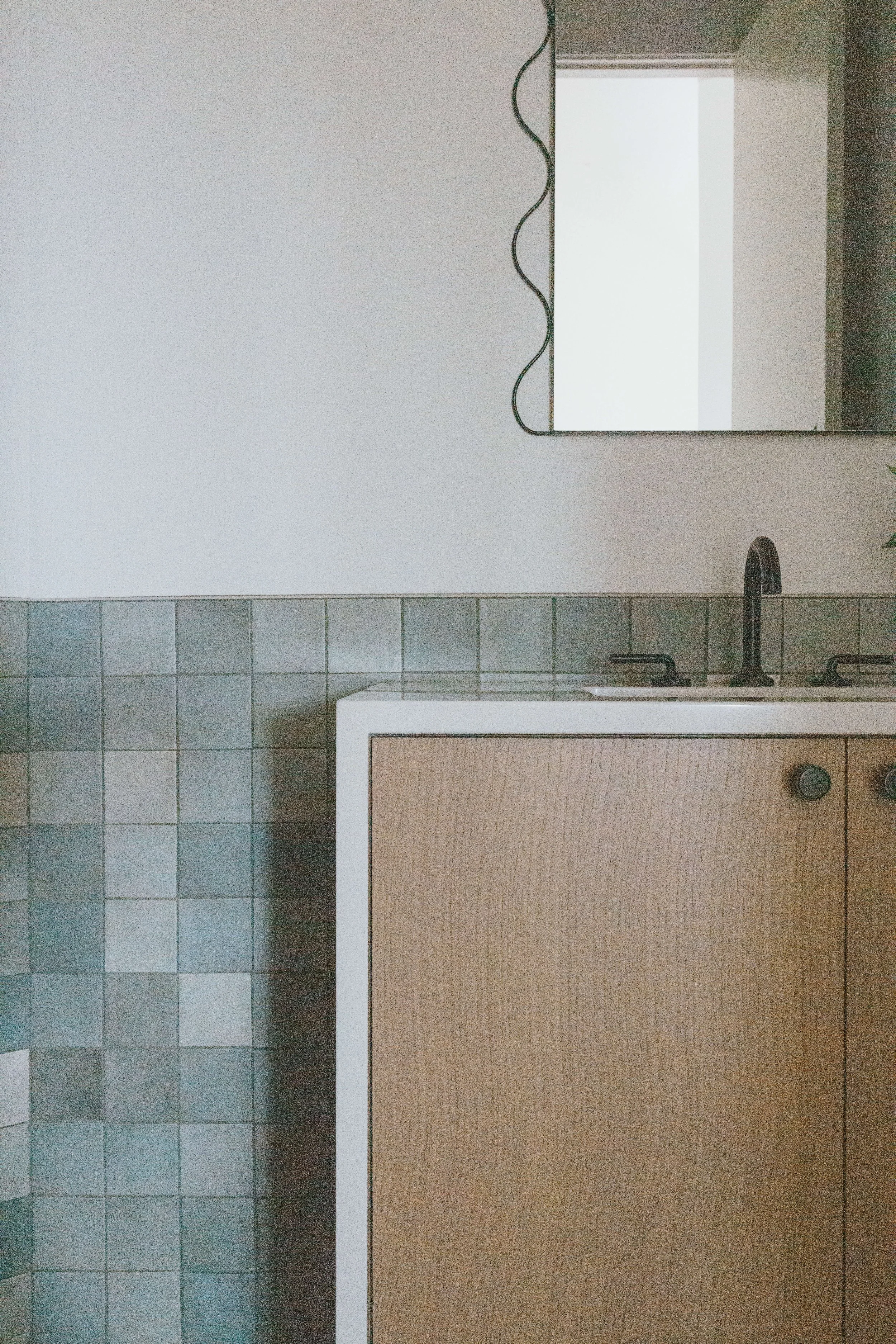 A bathroom with a white rectangular mirror above a light brown wooden cabinet with a black faucet. There are light gray tiles along the wall and darker tiles along the floor. An electrical cord is hanging from the ceiling.