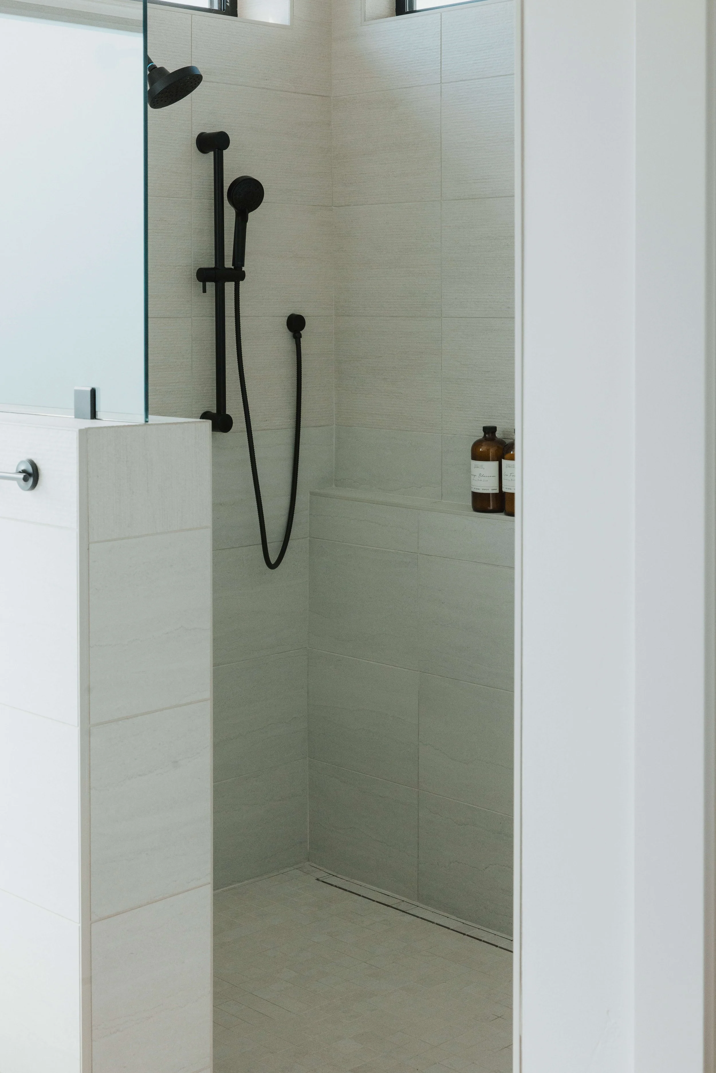 A modern shower with a dark showerhead and a movable hand-held shower in a corner, with bottles of shampoo or soap on a built-in shelf, and a small drain on the tiled floor.