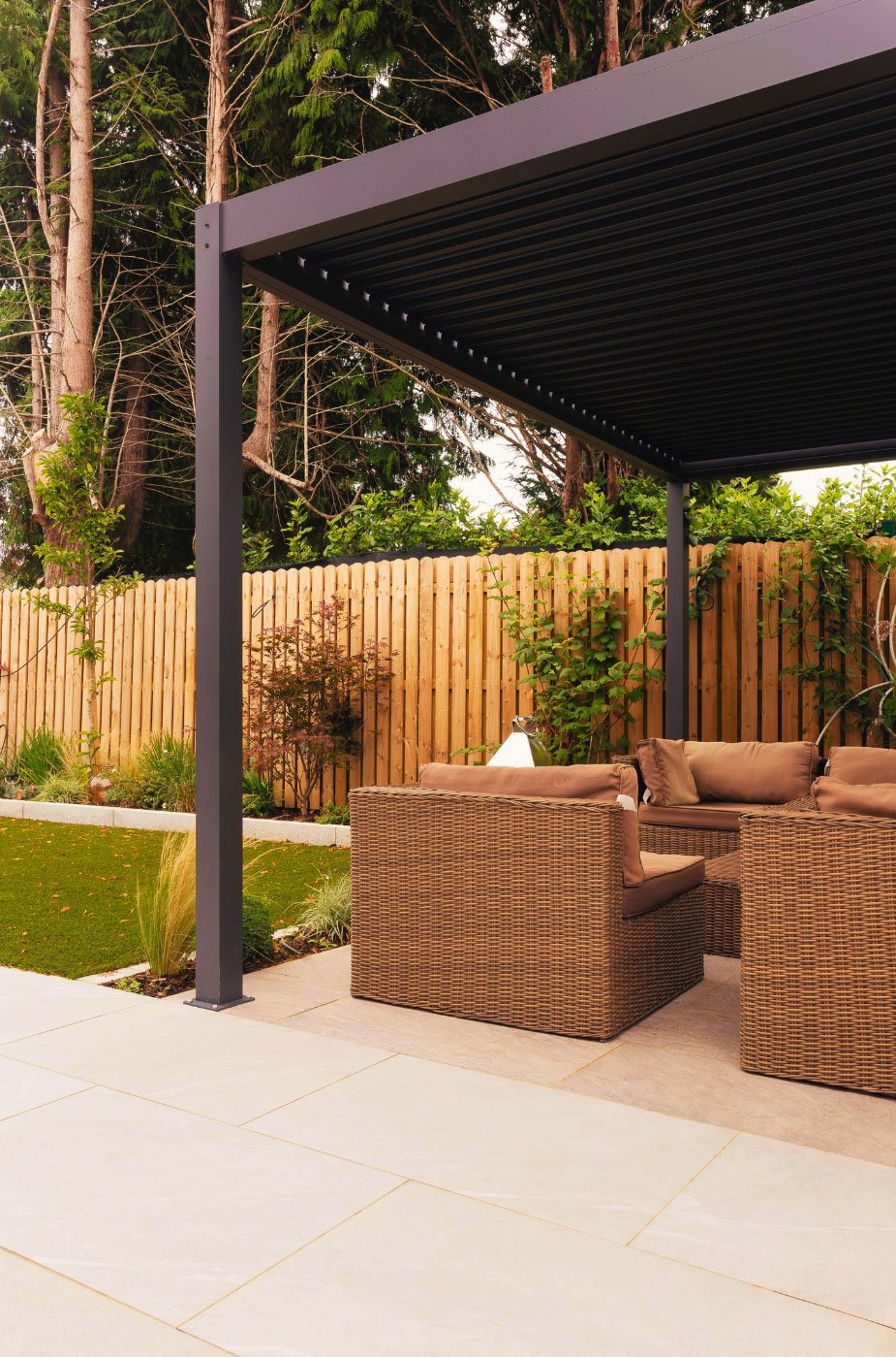 Outdoor patio with wicker furniture under a black metal pergola, wooden fence, and garden plants, with a background of trees.