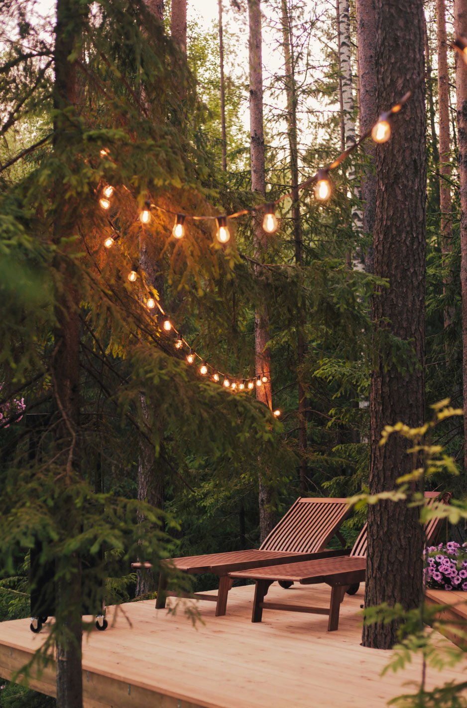 Wooden deck with lounge chair and string lights hanging among trees in a forest setting at sunset.