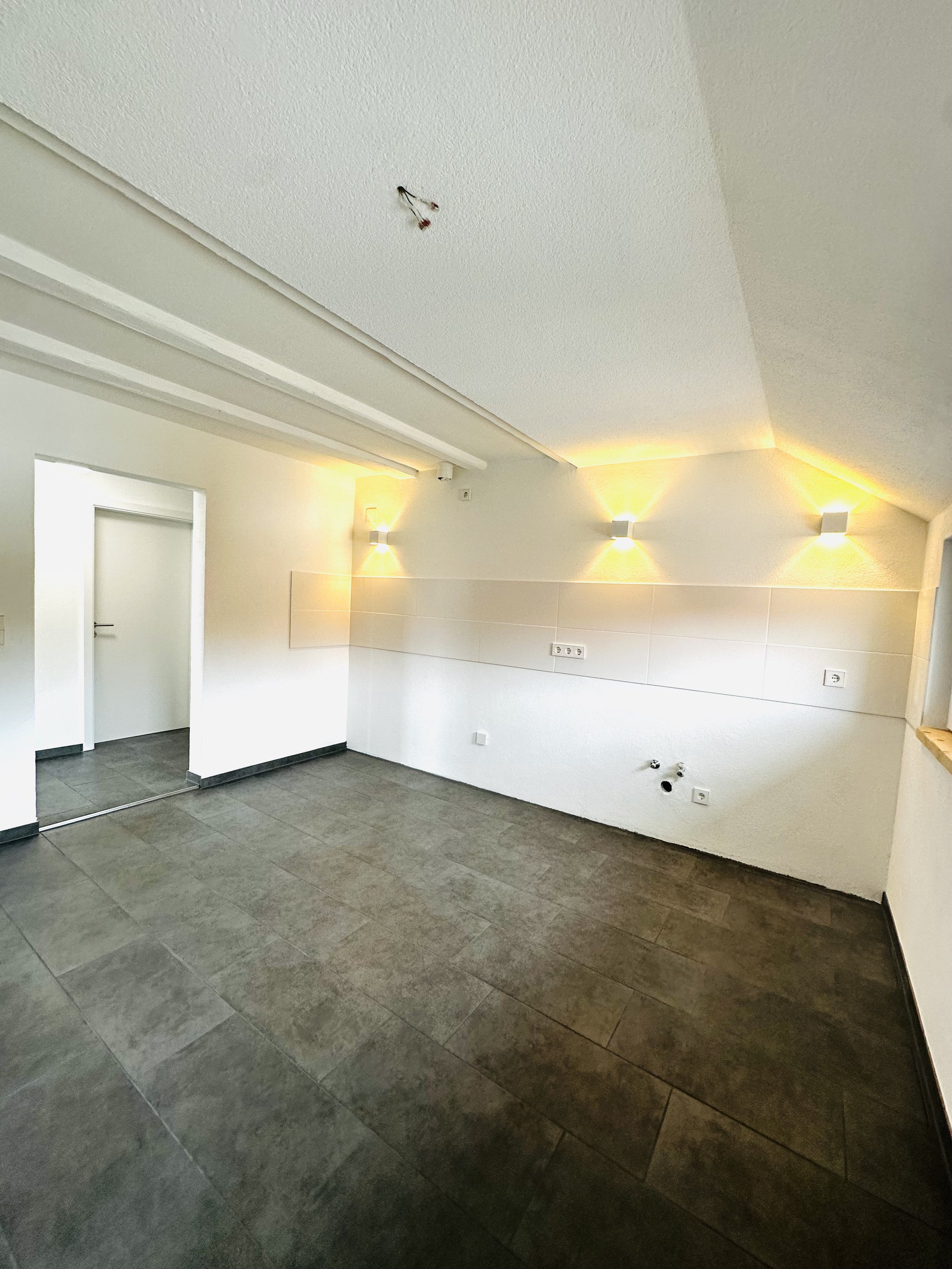 Leerer, moderner Raum mit dunklem Fliesenboden und weißen Wänden. Drei Wandlampen leuchten eine Wand aus. An der Decke hängen freiliegende Kabel. Es gibt Anschlüsse für eine Küche an der rechten Wand und eine offene Türöffnung links.