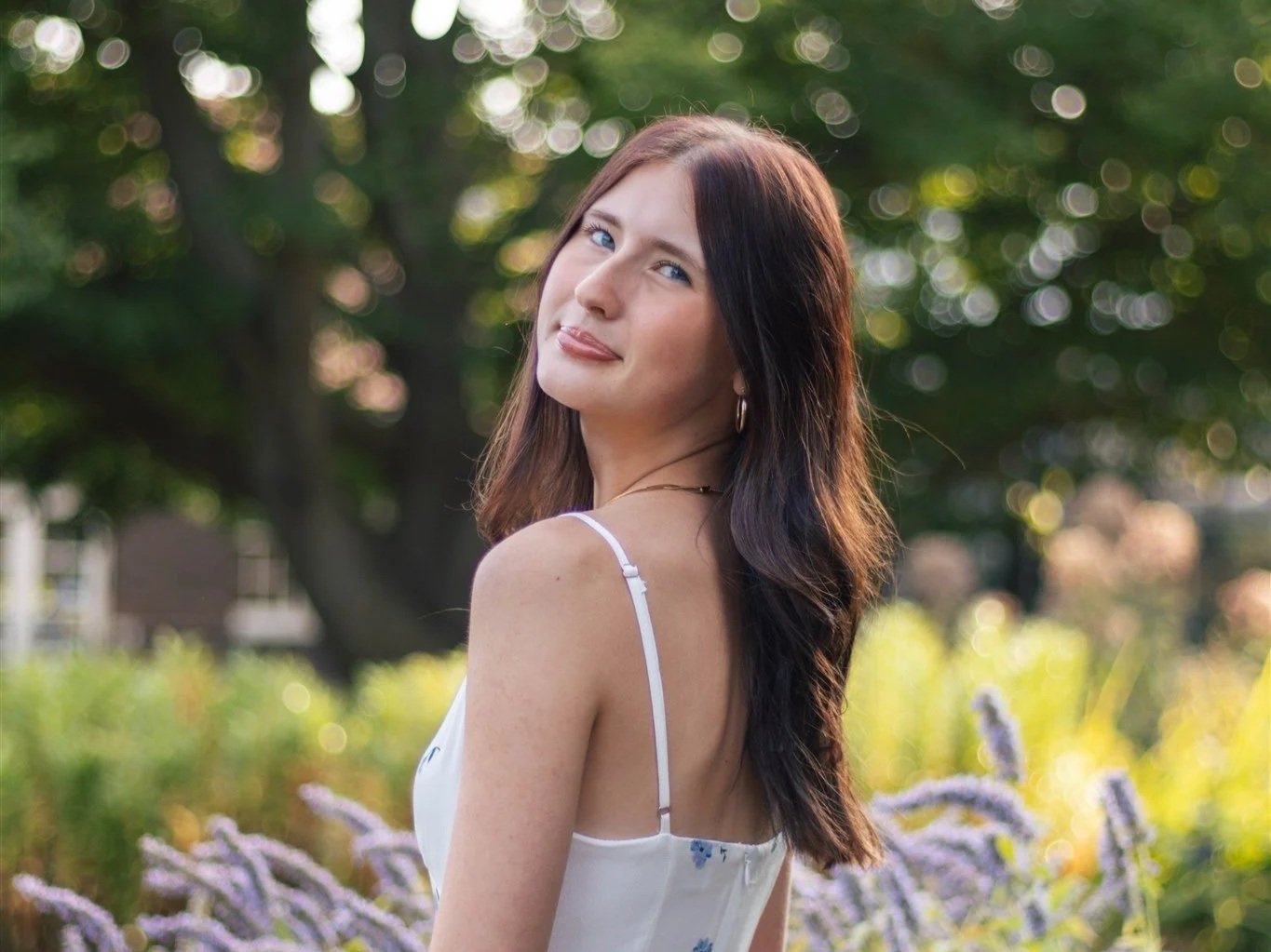 A woman with long brown hair and blue eyes is outdoors in a garden or park, wearing a white spaghetti strap top. She is looking over her shoulder towards the camera with a slight smile, surrounded by greenery and purple flowers.