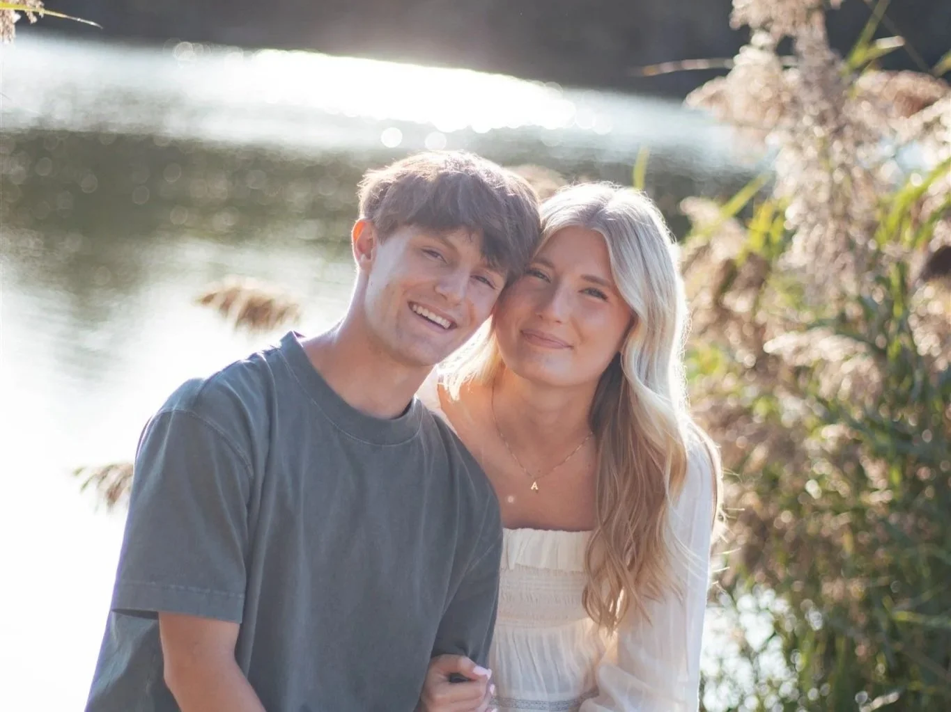 A young couple smiling outdoors near water with trees in the background, bathed in soft sunlight.