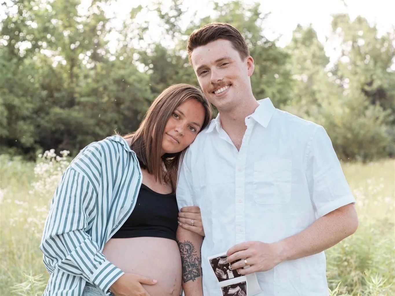 A young couple with a pregnant woman leaning her head on a man's shoulder outdoors, smiling at the camera, with a grassy field and trees in the background.