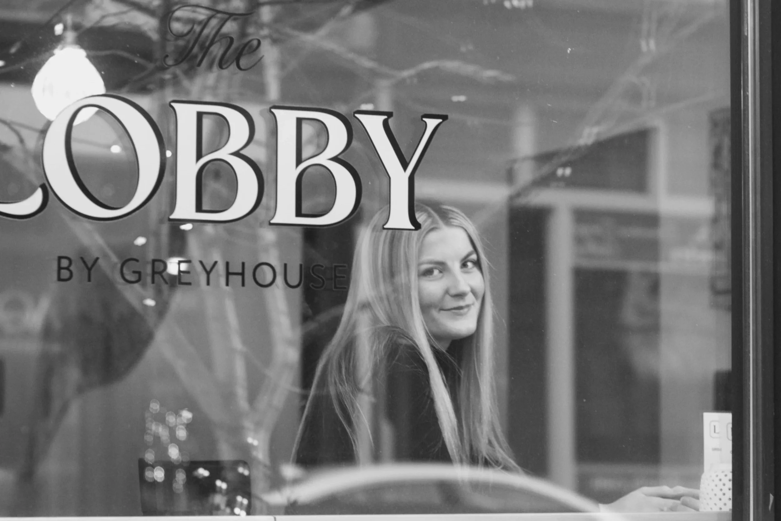 A woman with long blonde hair smiling through a retail store window with the words "The Lobby by Greyhouse" written on the glass.