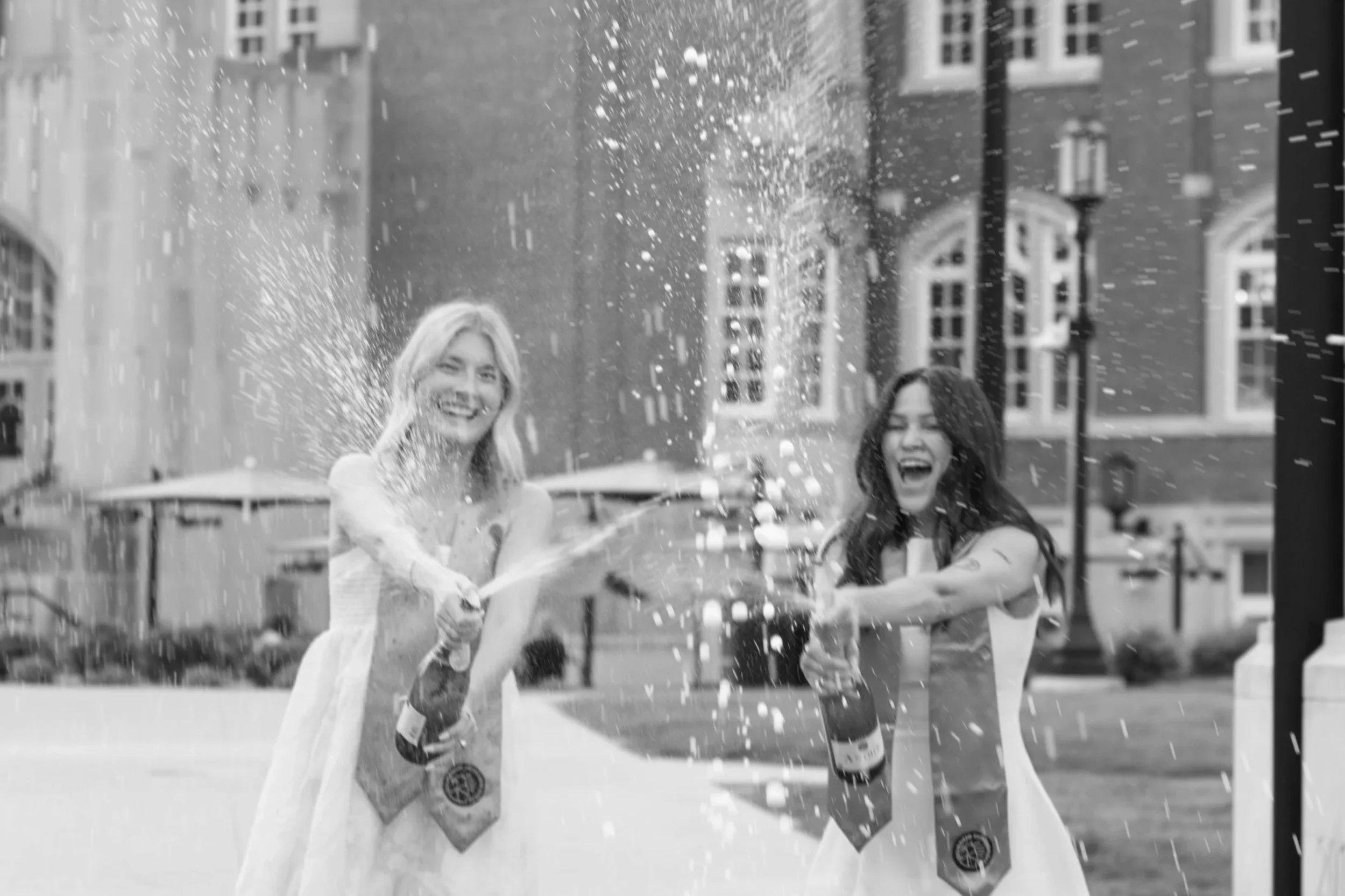 Two women in graduation gowns cheer and spray champagne outside on a city street.