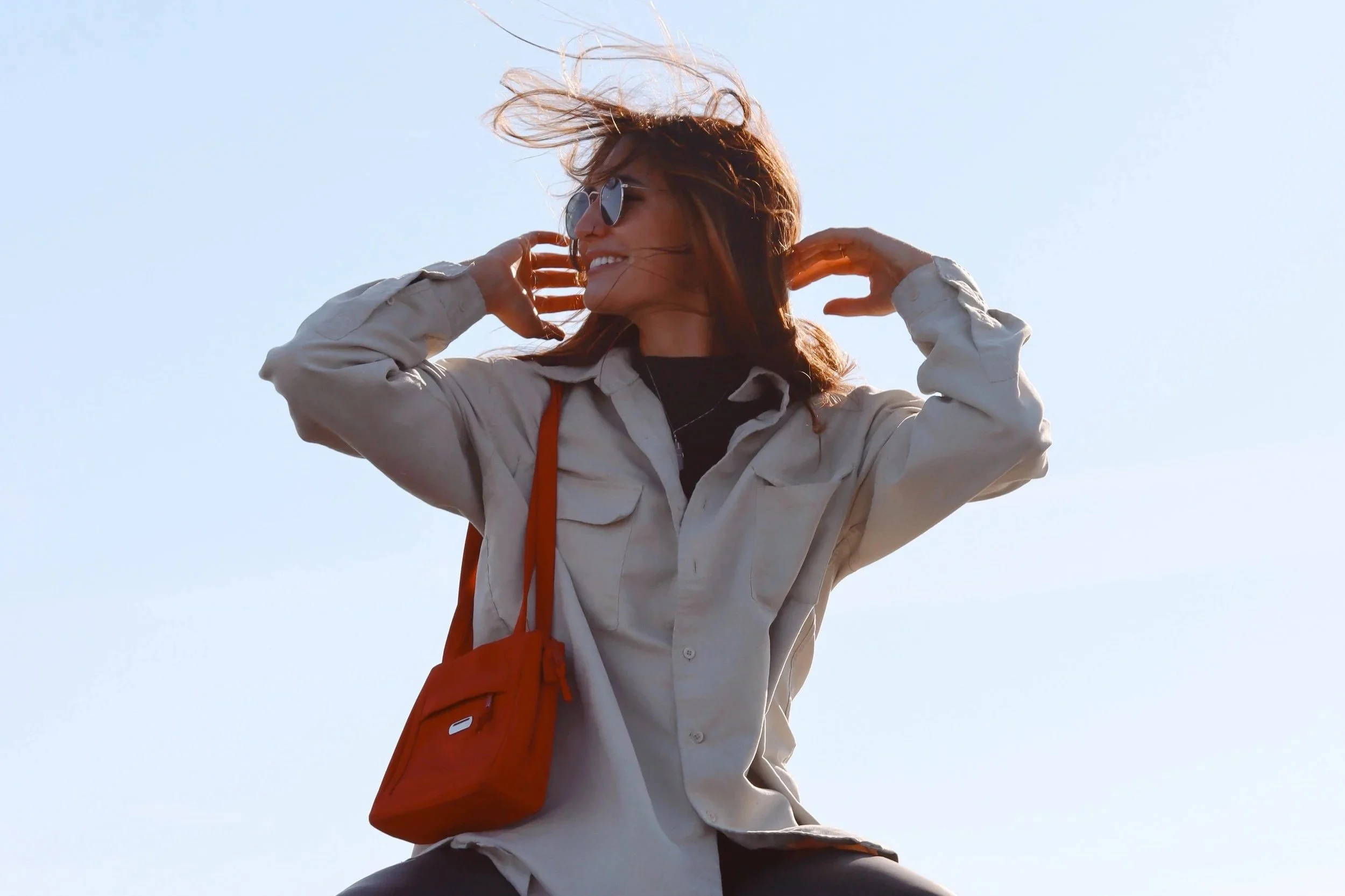 A woman with long hair wearing sunglasses, a beige jacket, and a black shirt, smiling and posing outdoors with a bright sky background. She has a red shoulder bag.