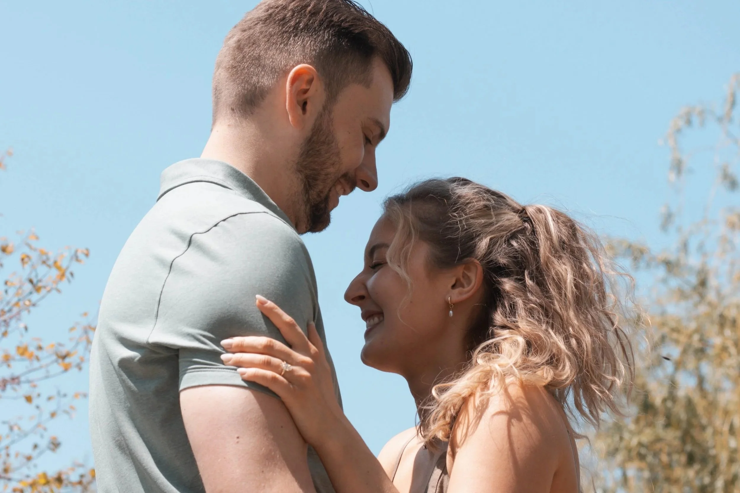 A happy couple, a man and a woman, embracing and smiling outdoors with blue sky and trees in the background.