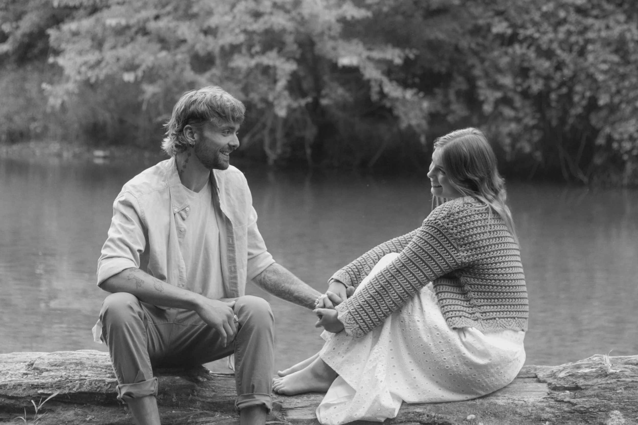 A man and woman sitting on a log by a lake, holding hands and smiling at each other in a black-and-white outdoor setting.