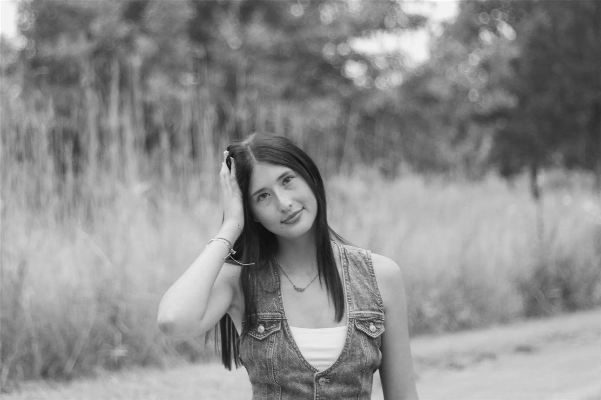 A young woman with long dark hair, wearing a sleeveless denim vest and a white top, standing outdoors in a grassy area with trees in the background. She has her hand on her head and is smiling gently at the camera.