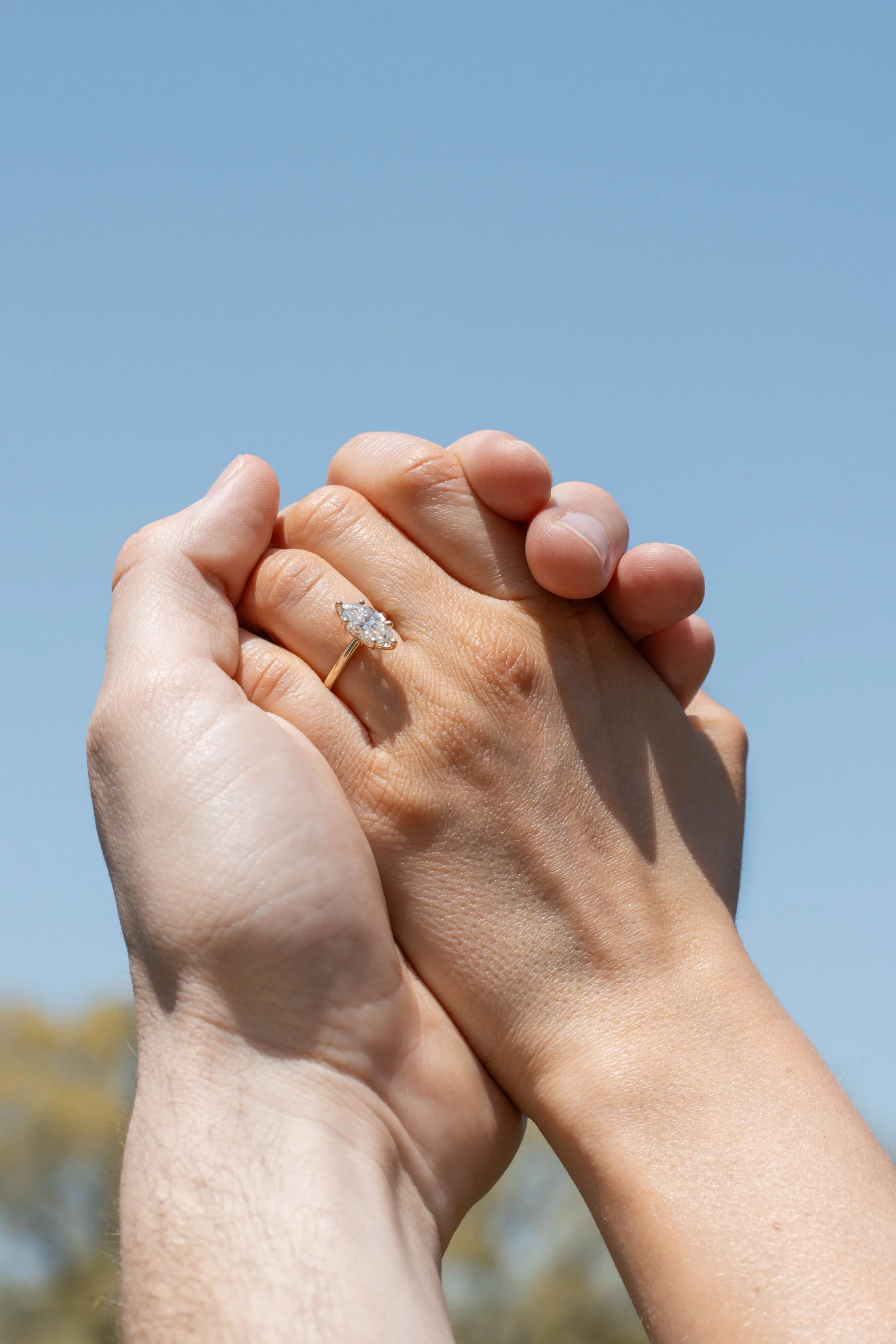 Two hands clasping, one wearing a diamond engagement ring, against a clear blue sky.