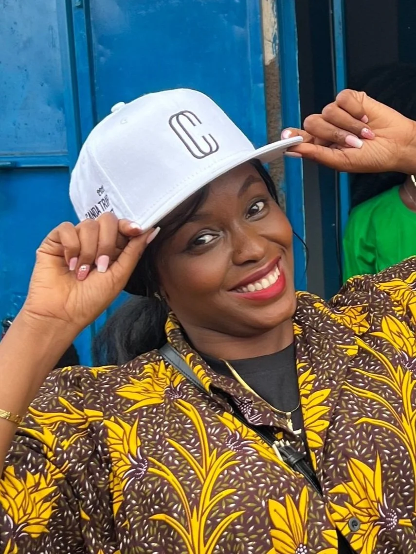 A woman smiling, wearing a patterned yellow and brown shirt, and a white baseball cap with a logo, tilting the hat back with her hands, standing in front of a blue background.