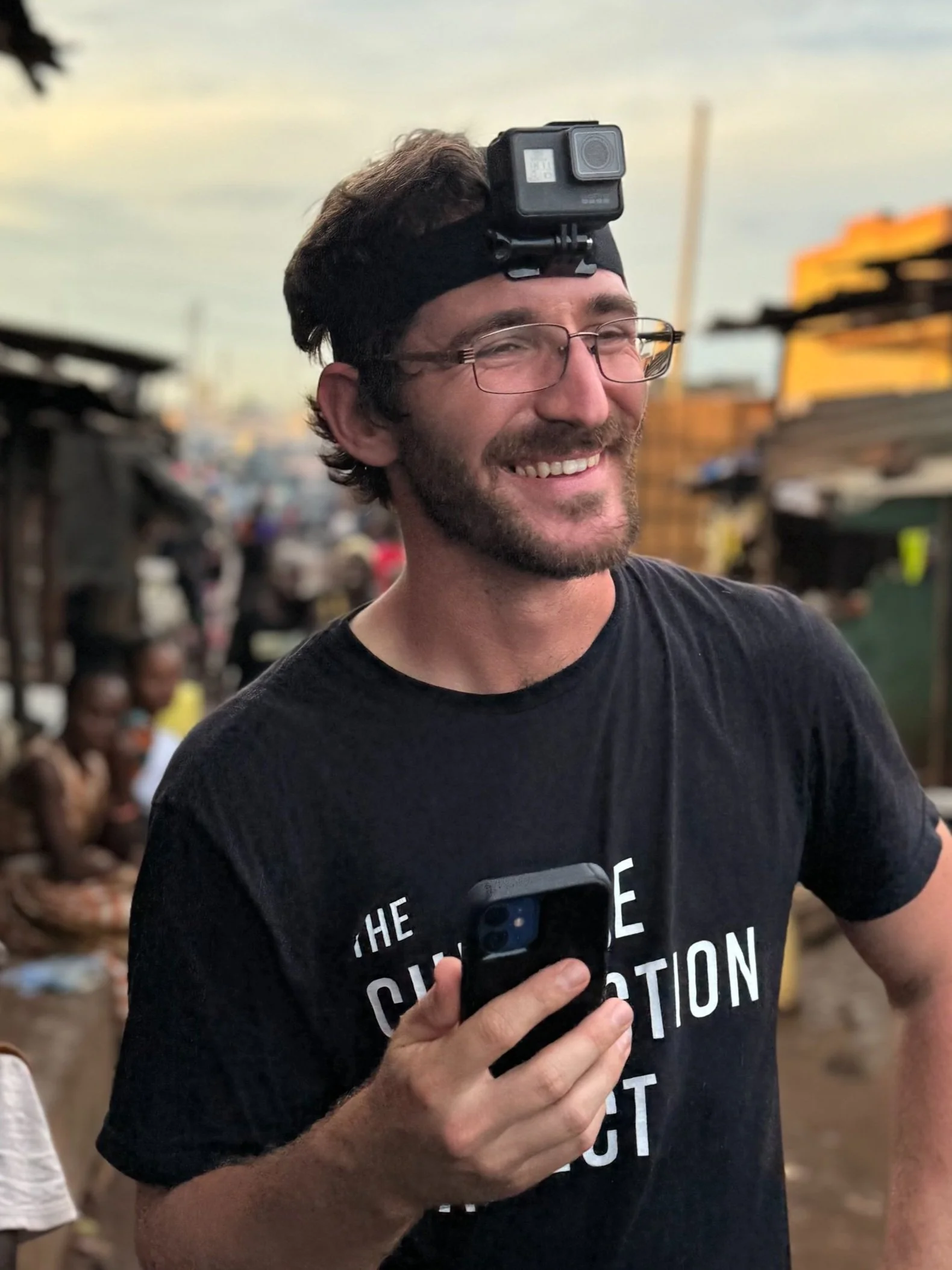 A smiling man with a beard, glasses, and dark hair wearing a black T-shirt that says 'The Challenge of Creation.' He is holding a smartphone in his right hand and has a GoPro camera mounted on a head strap, standing outdoors with a blurred background of a marketplace with children and market stalls.
