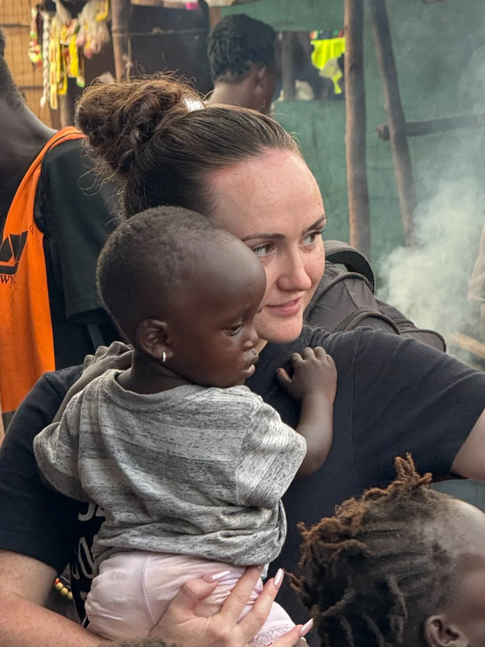 A woman with dark hair pulled back in a bun holding a young African girl in her arms. The girl has earrings, short hair, and is wearing a gray shirt. They are in a setting with other people and visible smoke or steam in the background.