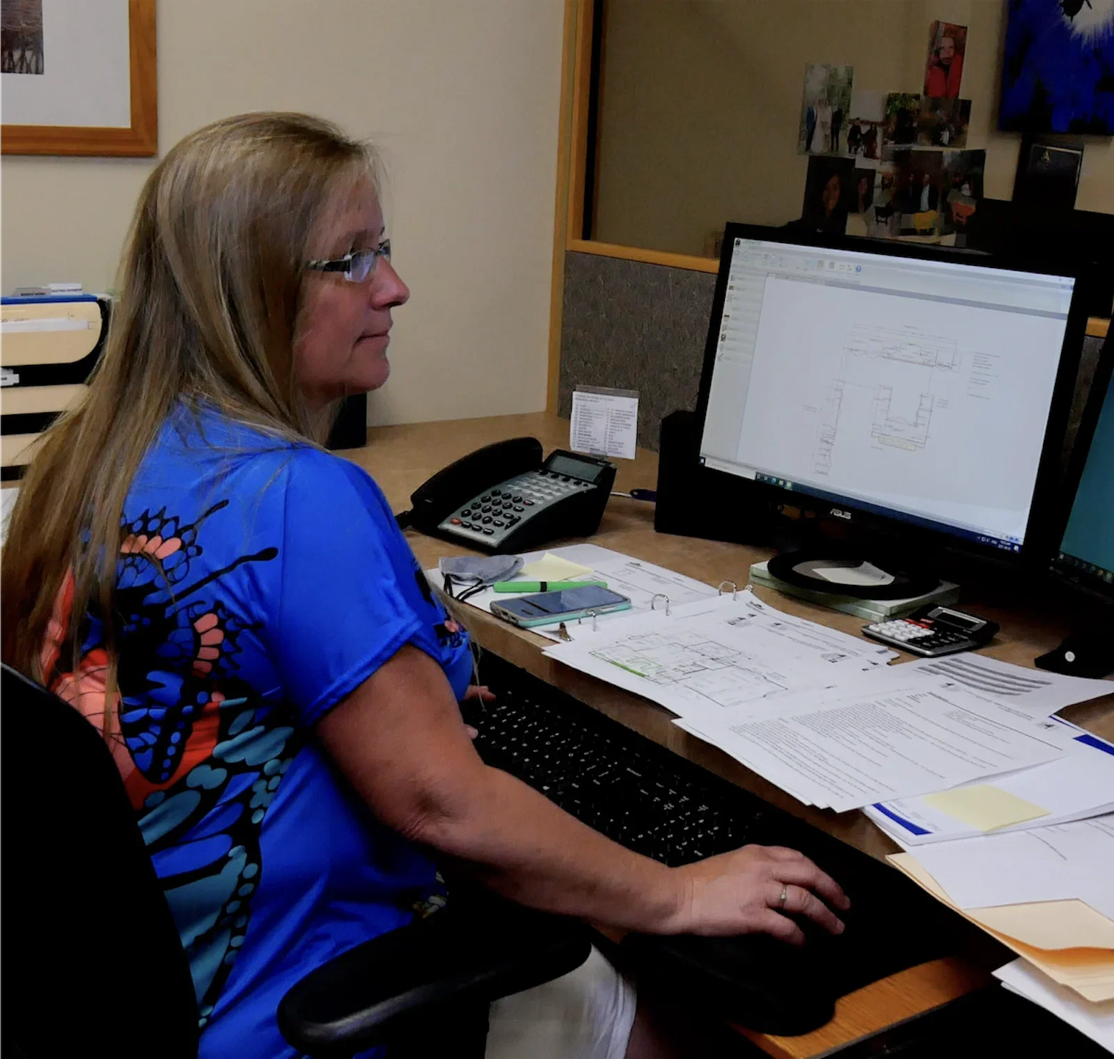 Woman with glasses working at a computer desk with a desktop monitor, phone, and papers.