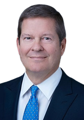 Headshot of a middle-aged man with short brown hair, wearing a dark suit, white shirt, and a blue tie, smiling against a plain white background.