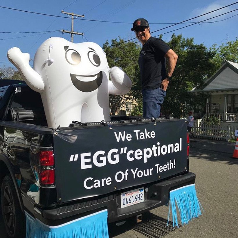 Man standing in a pickup truck bed next to an inflatable tooth; banner reads ‘We Take “EGG”ceptional Care Of Your Teeth!"