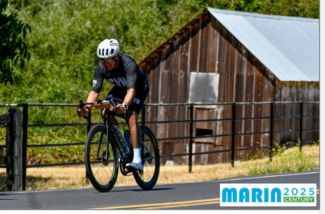 Cyclist riding on a road near a barn; ‘MARIN 2025 CENTURY’ logo in the corner