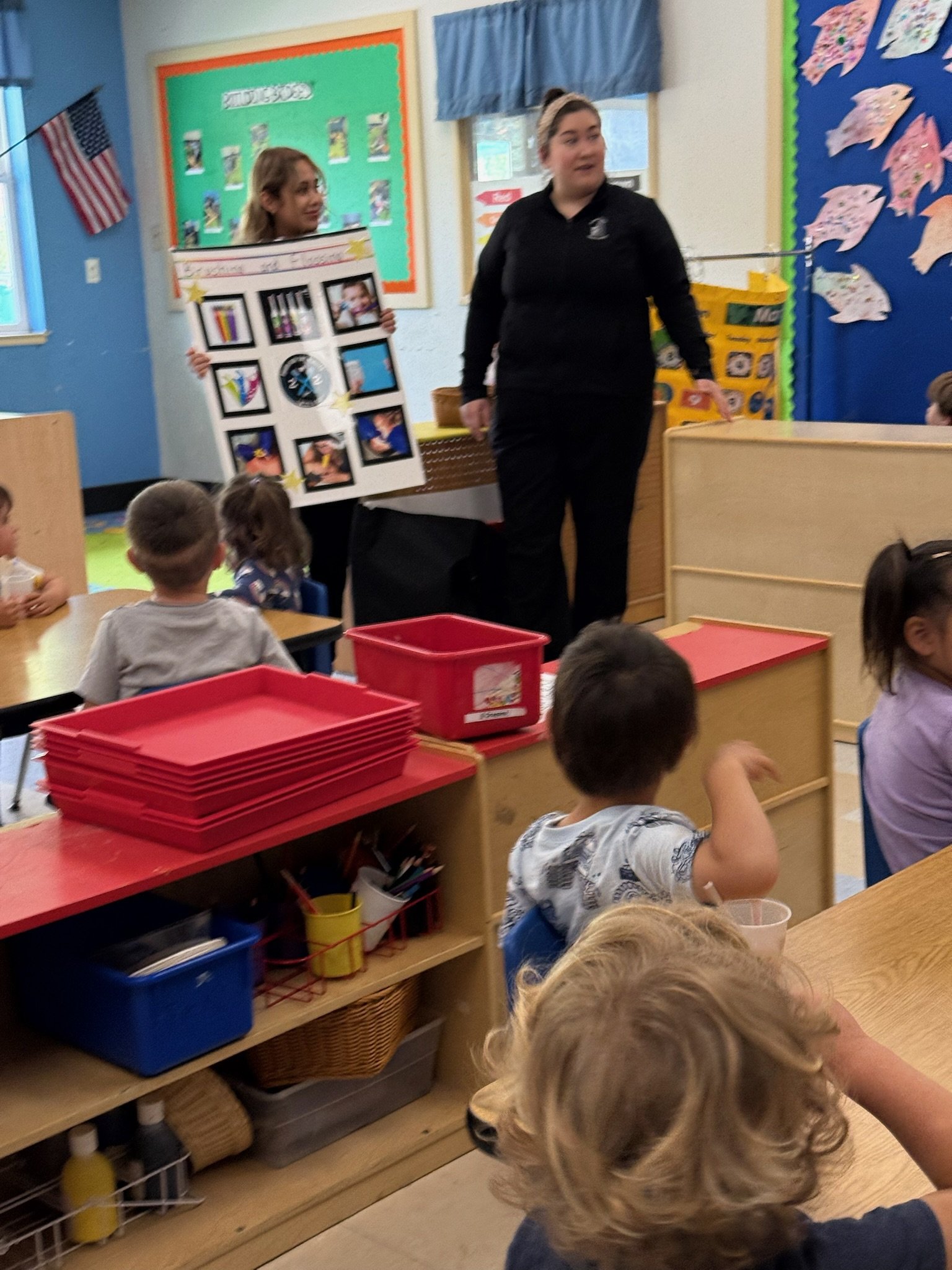 Two adults presenting to a classroom of young children; one holds a poster with photo examples